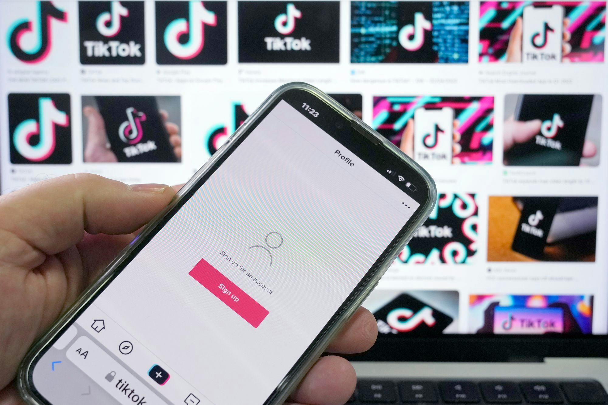 A sign up page for the application TikTok is shown on a cell phone in front of a screen with logos for the company.