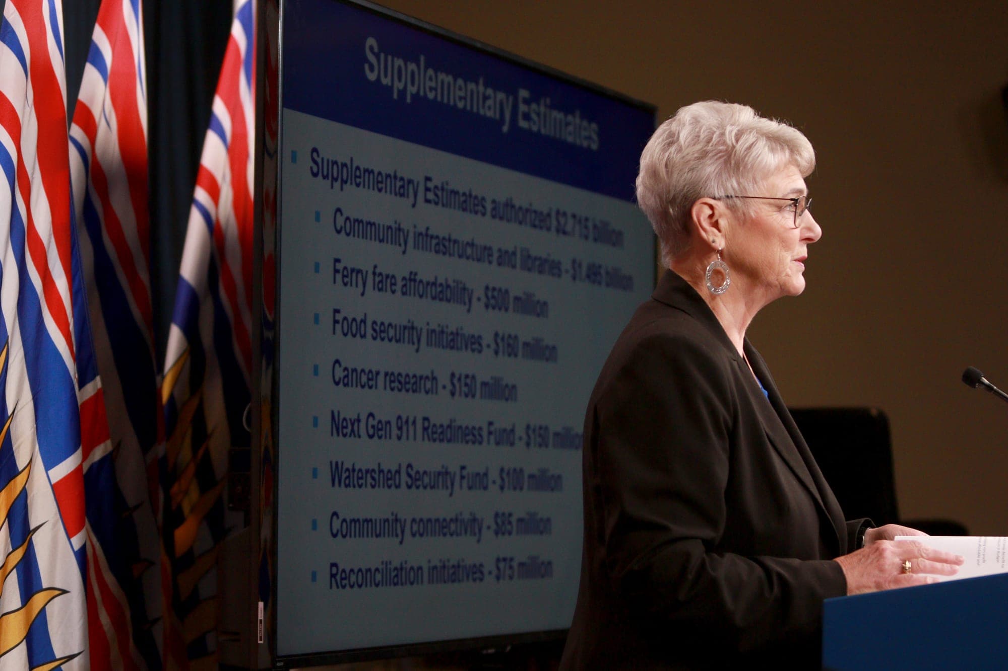 A side profile of woman wearing glasses and a black suit standing at a podium. 