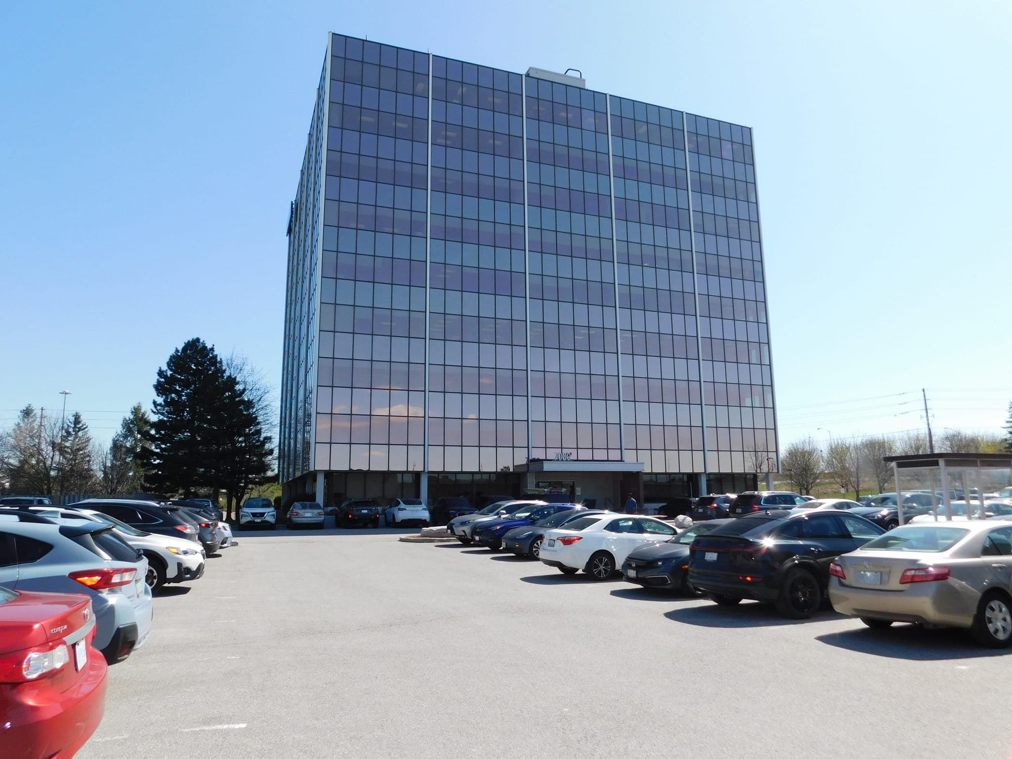 A glass office tower is pictured behind a concrete parking lot full of cars. 