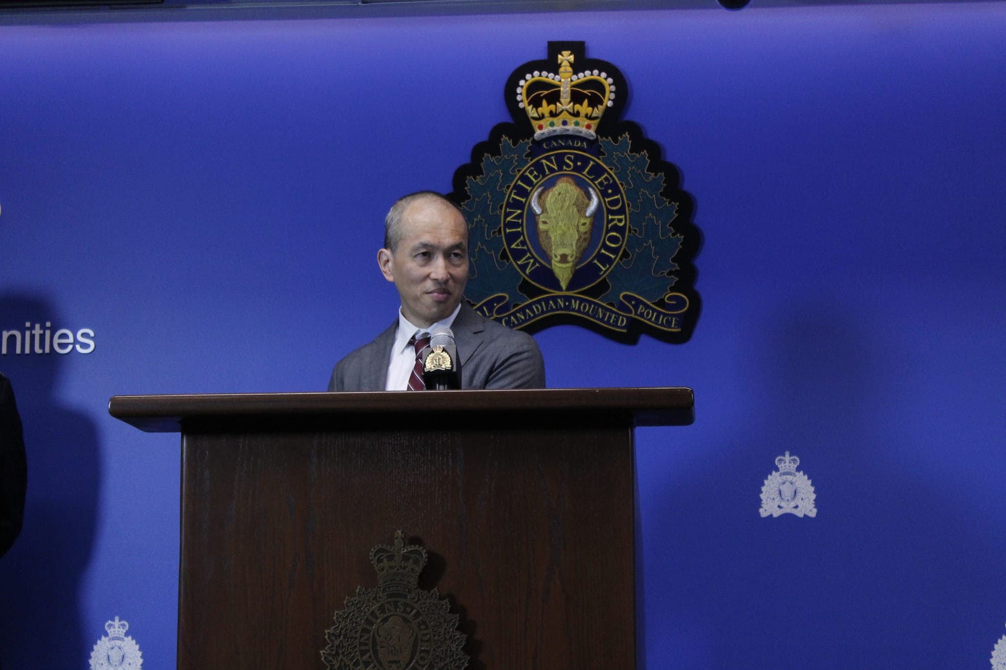 A man in a grey suite stands behind a podium. There is a dark blue background bearing the sigil of the BC Royal Canadian Mounted Police. 