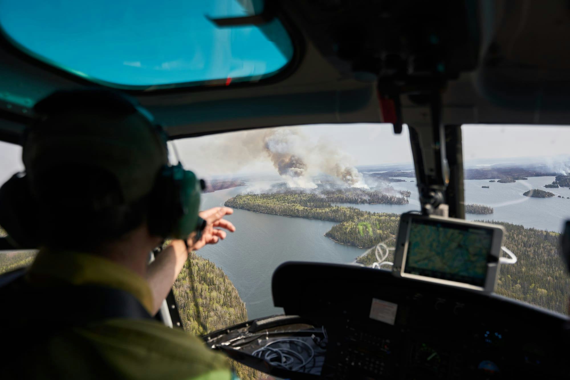 A person inside a helicopter gesturing to a forest in the distance that is billowing heavy smoke.