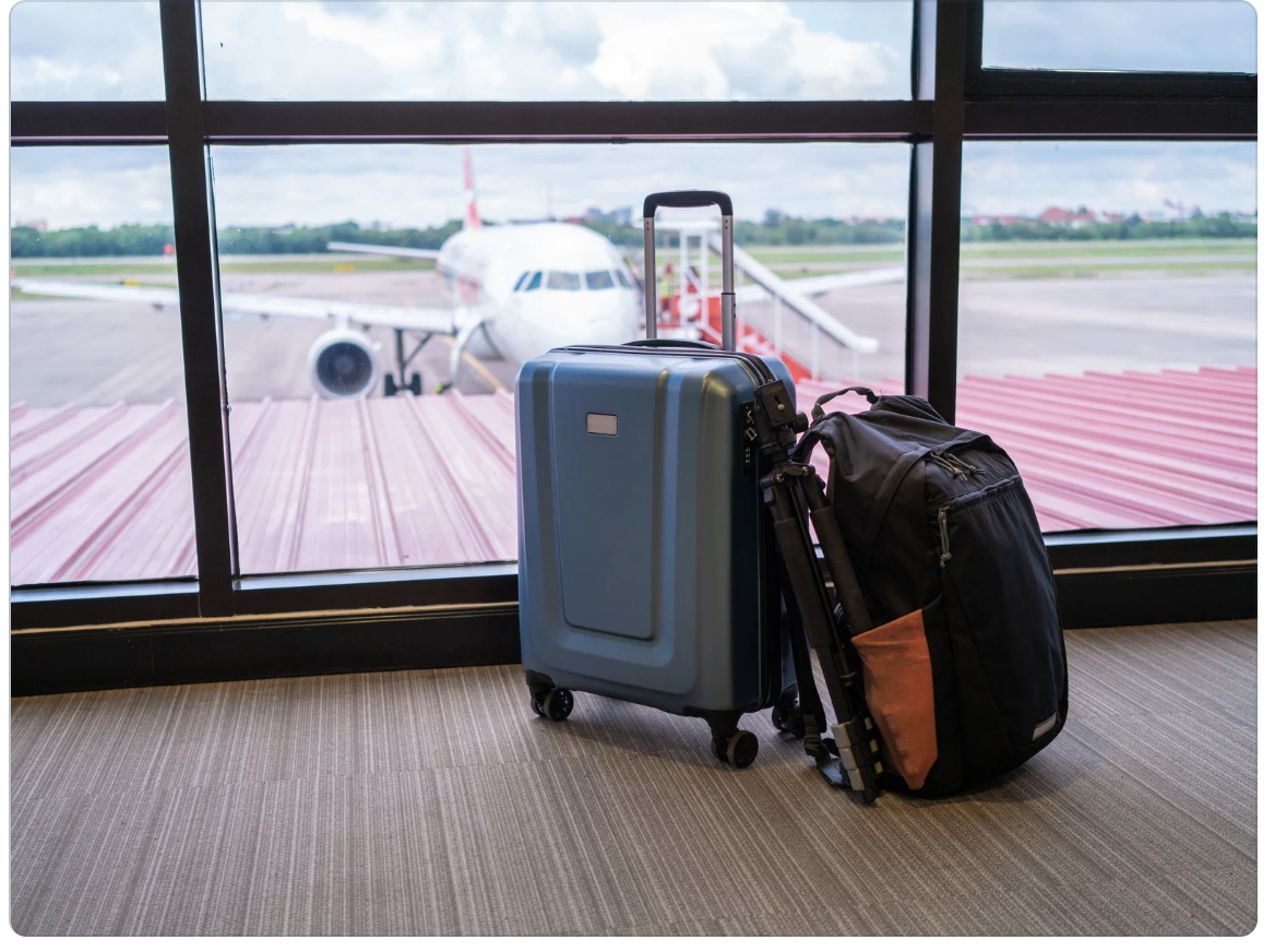 A backpack leans against a carry-on suitcase. Through a window behind the luggage, a plane can be seen parked on a tarmac. 