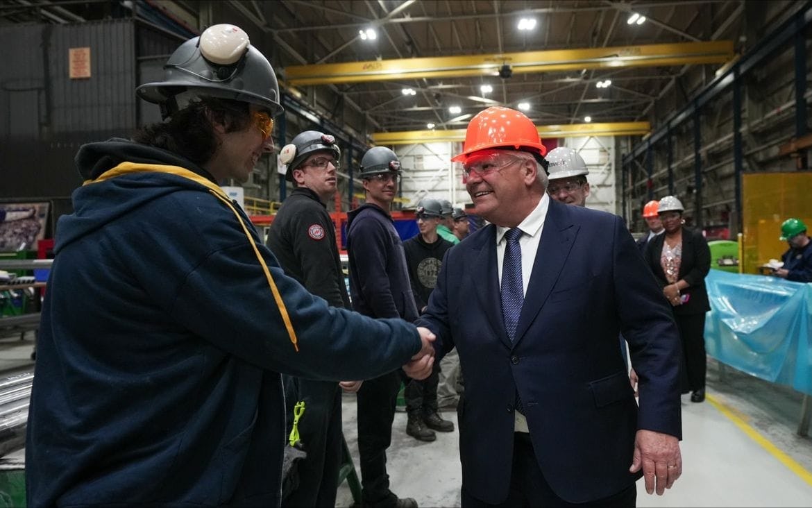 Doug Ford smiles and shakes the hand of a nuclear manufacturing plant worker while other workers look on in the background.