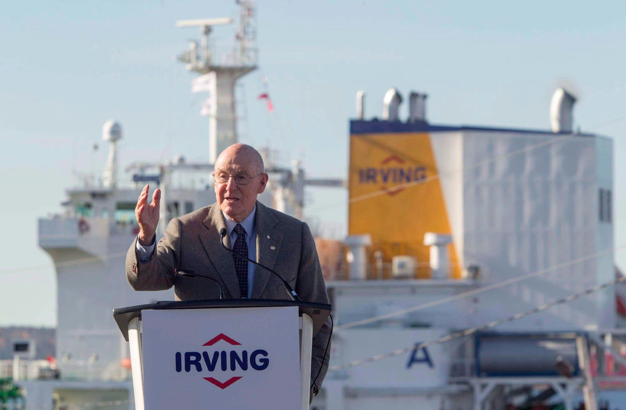 The former Irving stands behind a podium featuring the Irving Oil logo, with a large ship in the background.