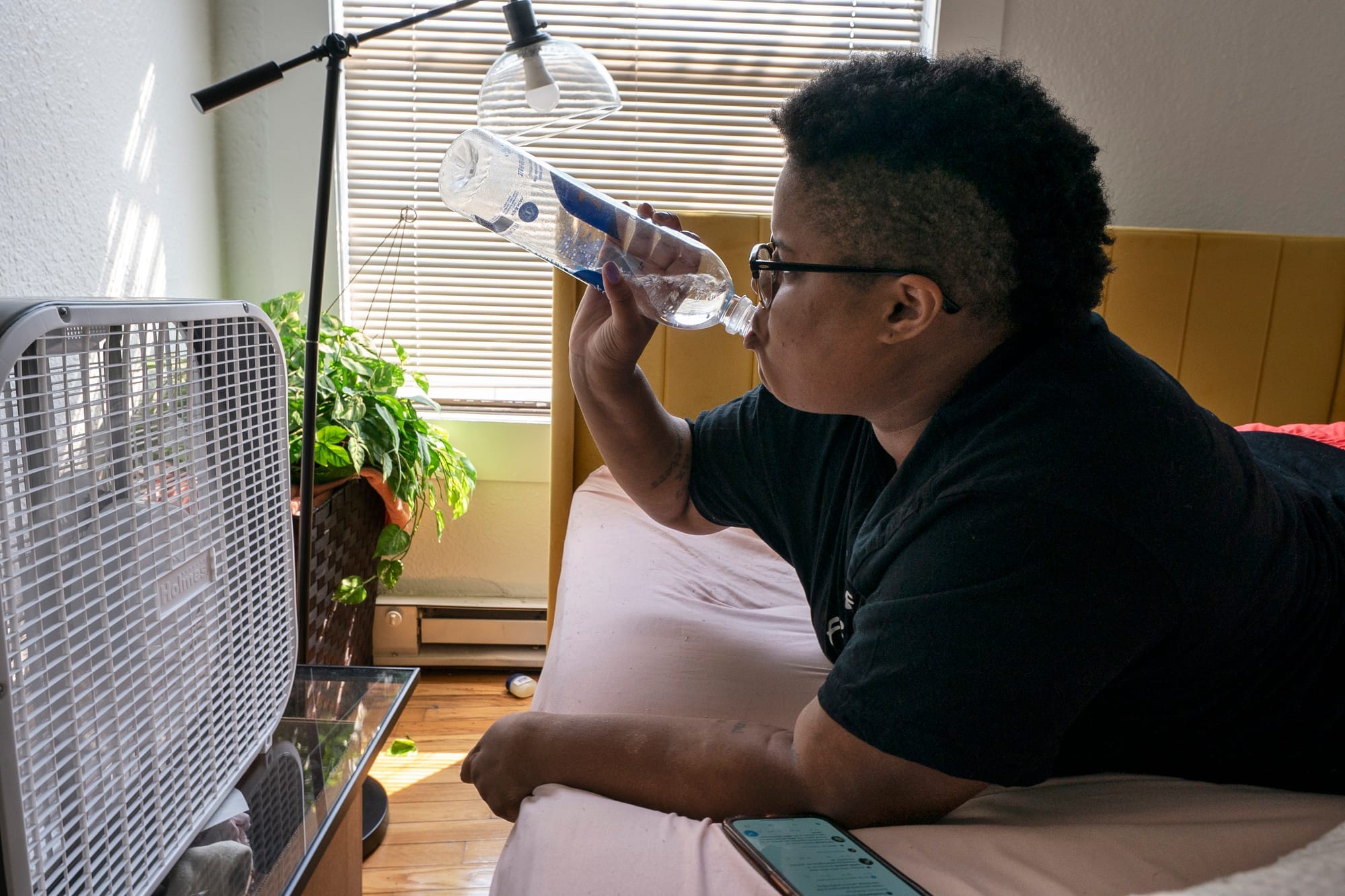 Someone rests on a bed, in front of a portable air-conditioning unit, and takes a sip of bottled water.