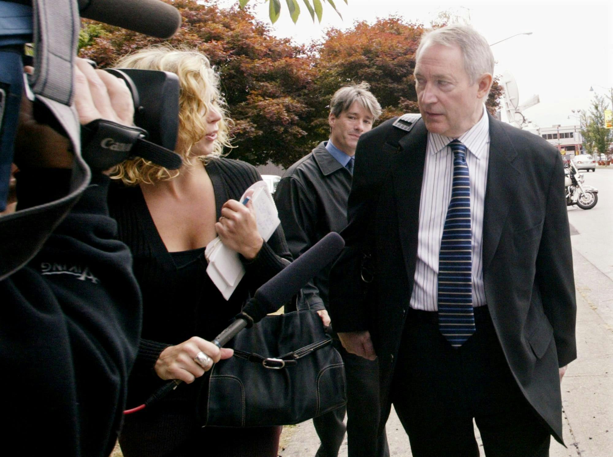 Michael Bolton, dressed in a black suit and striped tie, speaks to reporters on a sidewalk. 