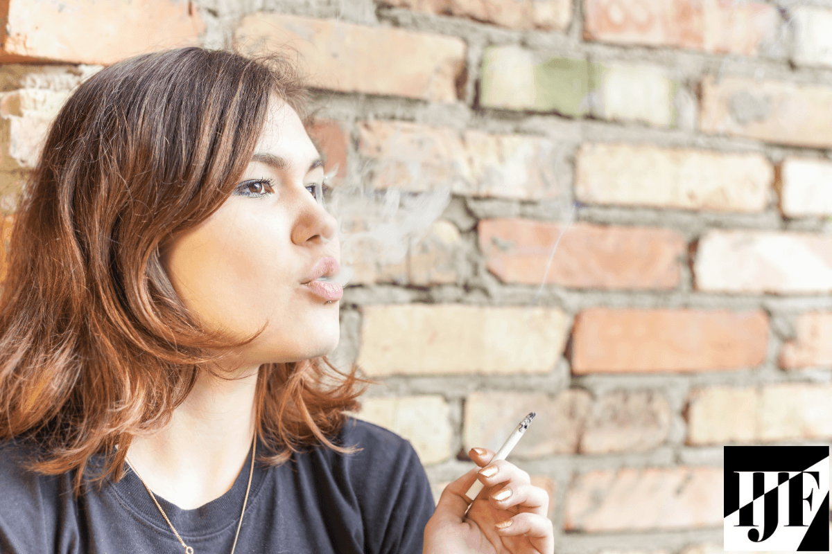 A young girl is blowing smoke from a cigarette in front of a brick wall. 