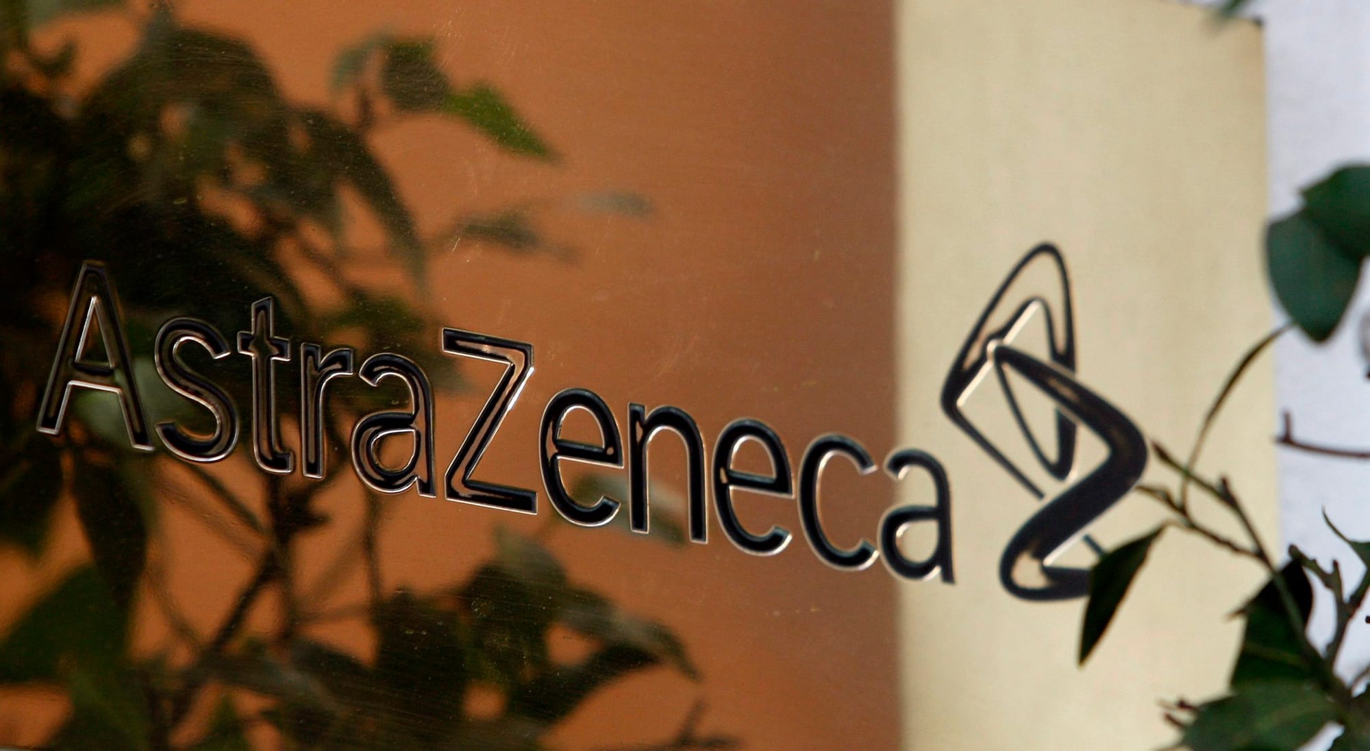 AstraZeneca's logo is shown in close-up on a mirrored surface, with green plants in the reflection.