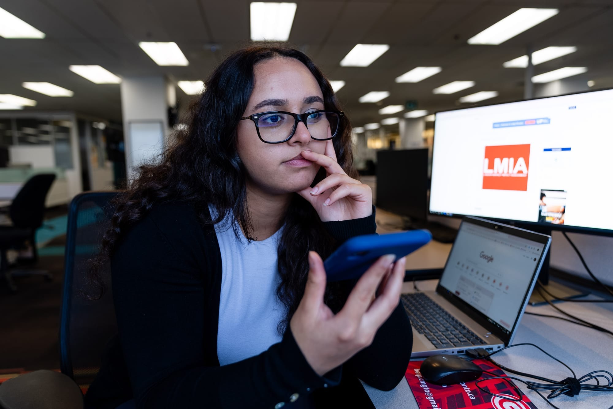 A young woman holds a blue cell phone, while a screen in the background shows an advertisement for an LMIA.