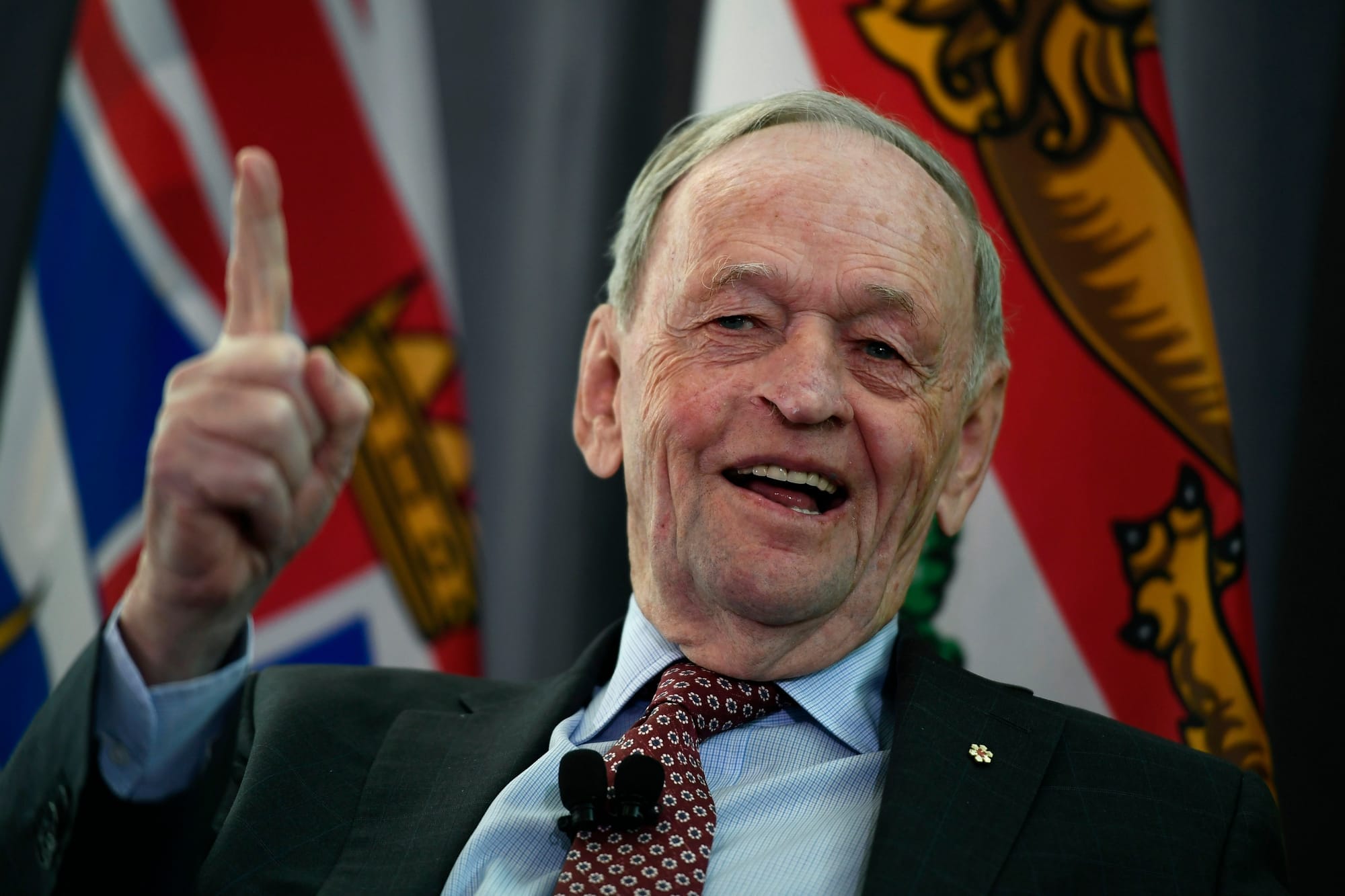 A man in his late 80s is wearing a suit jacket and tie as he smiles and speaks to a crowd