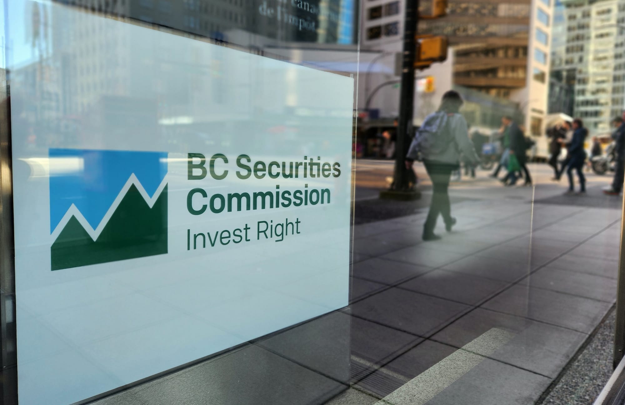 Pedestrians are reflected in a window where a sign reads "BC Securities Commission: Invest Right."