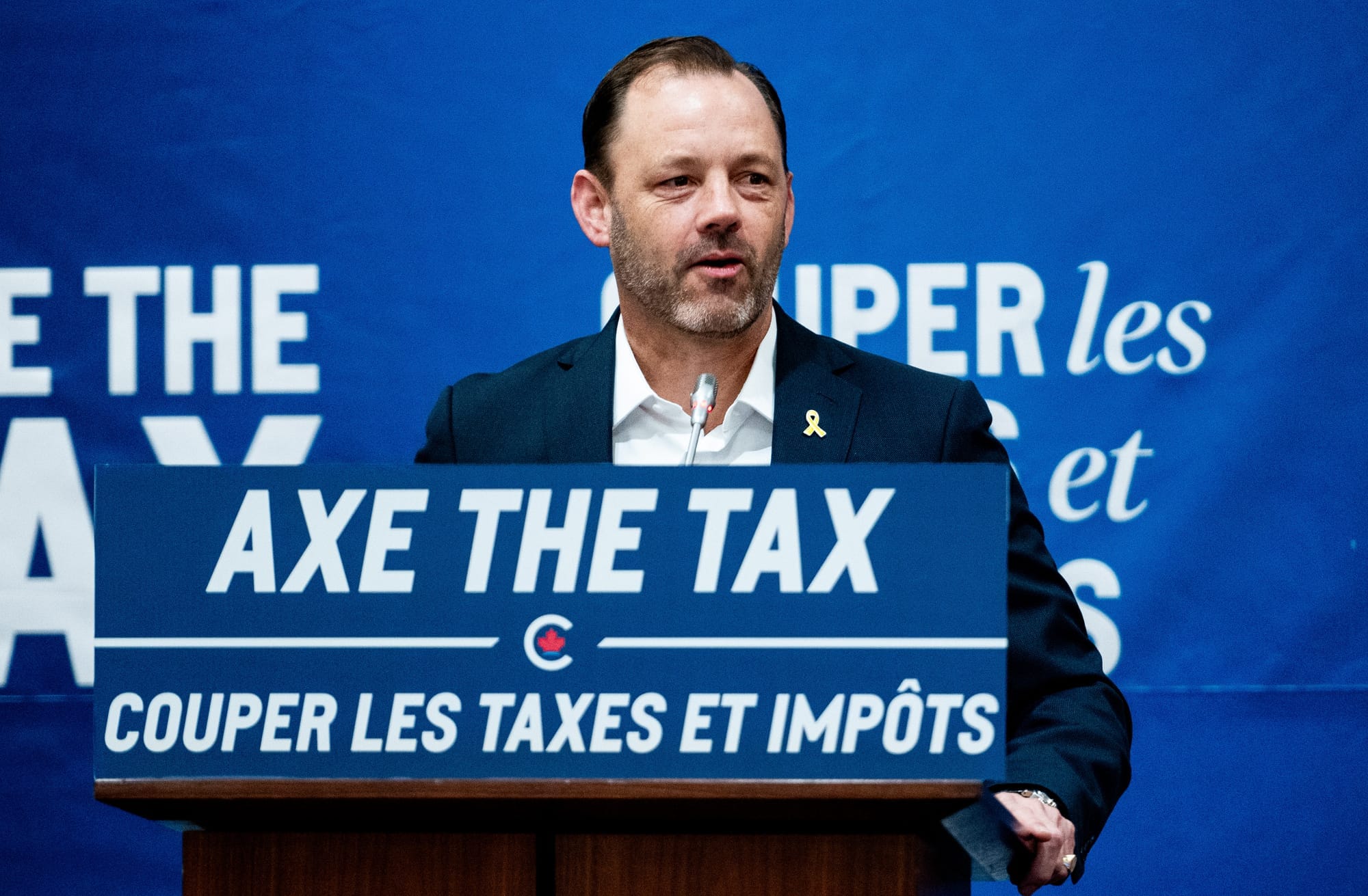 Don Stewart stands speaking behind a podium with an "Axe the Tax" Conservative-branded sign.