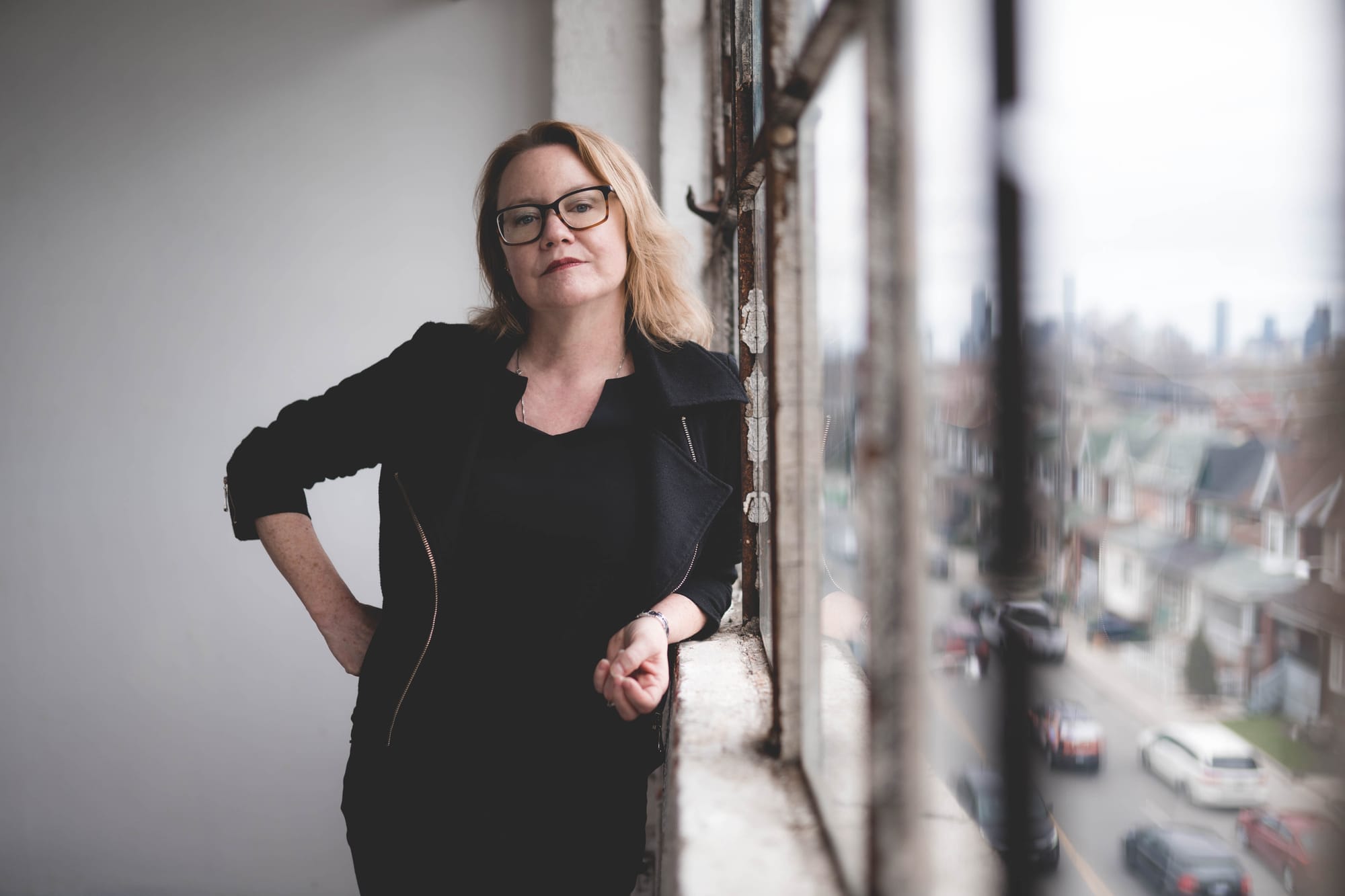 Shirley Moore wears a black outfit and leans against a window overlooking a city street.