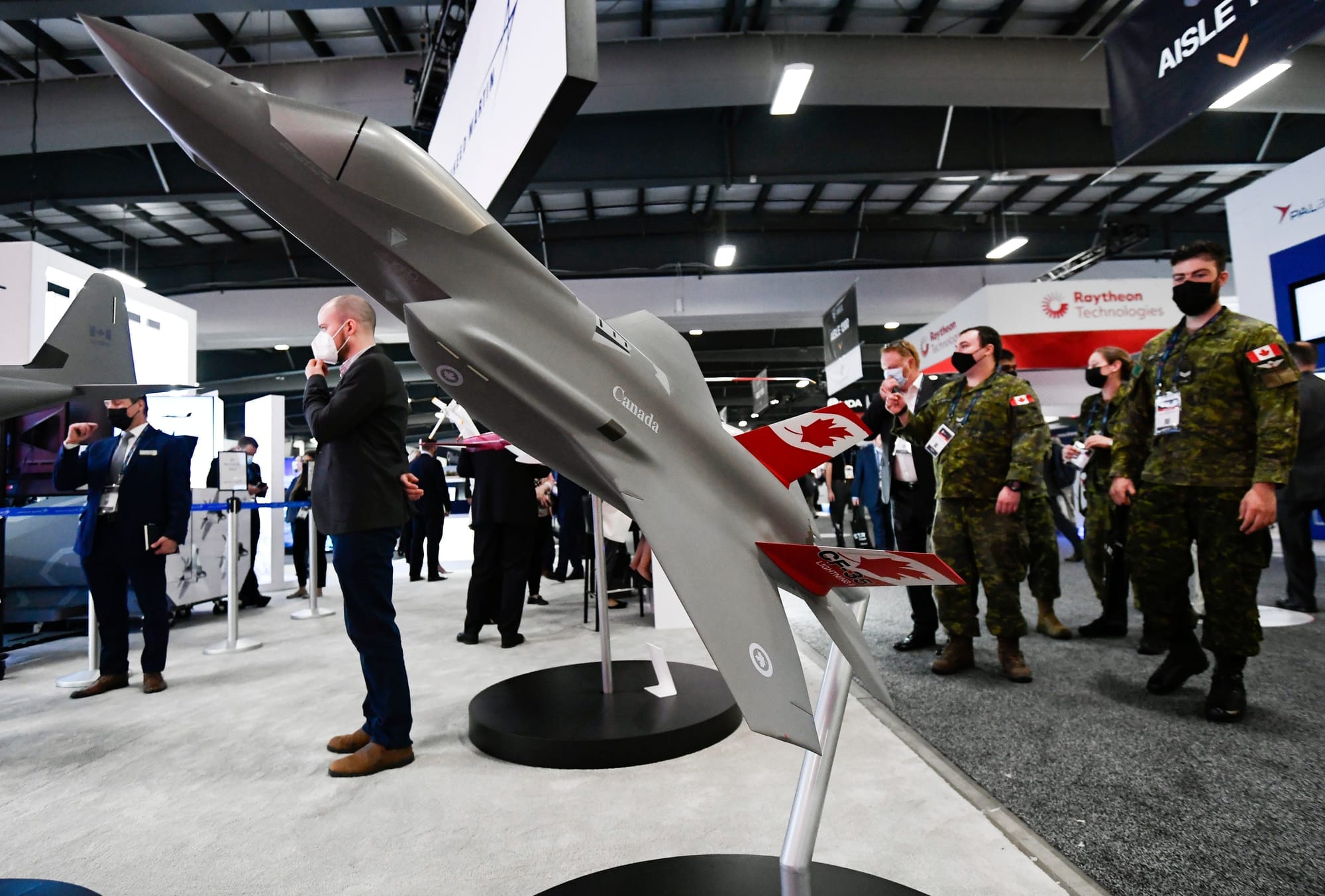 A model of a grey fighter jet is pictured in front of a group of people in suits and some in Canadian military uniforms. 