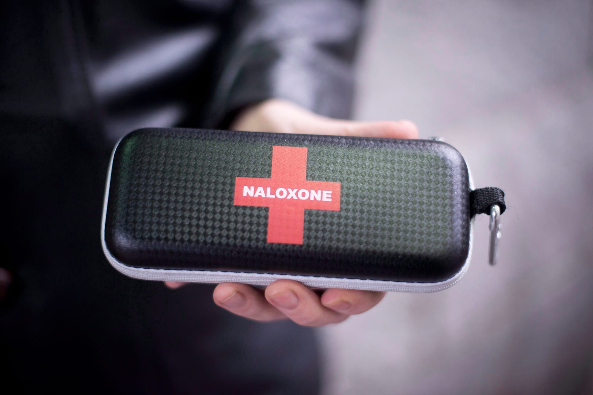 A person holds a black naloxone kit with a red cross on it.