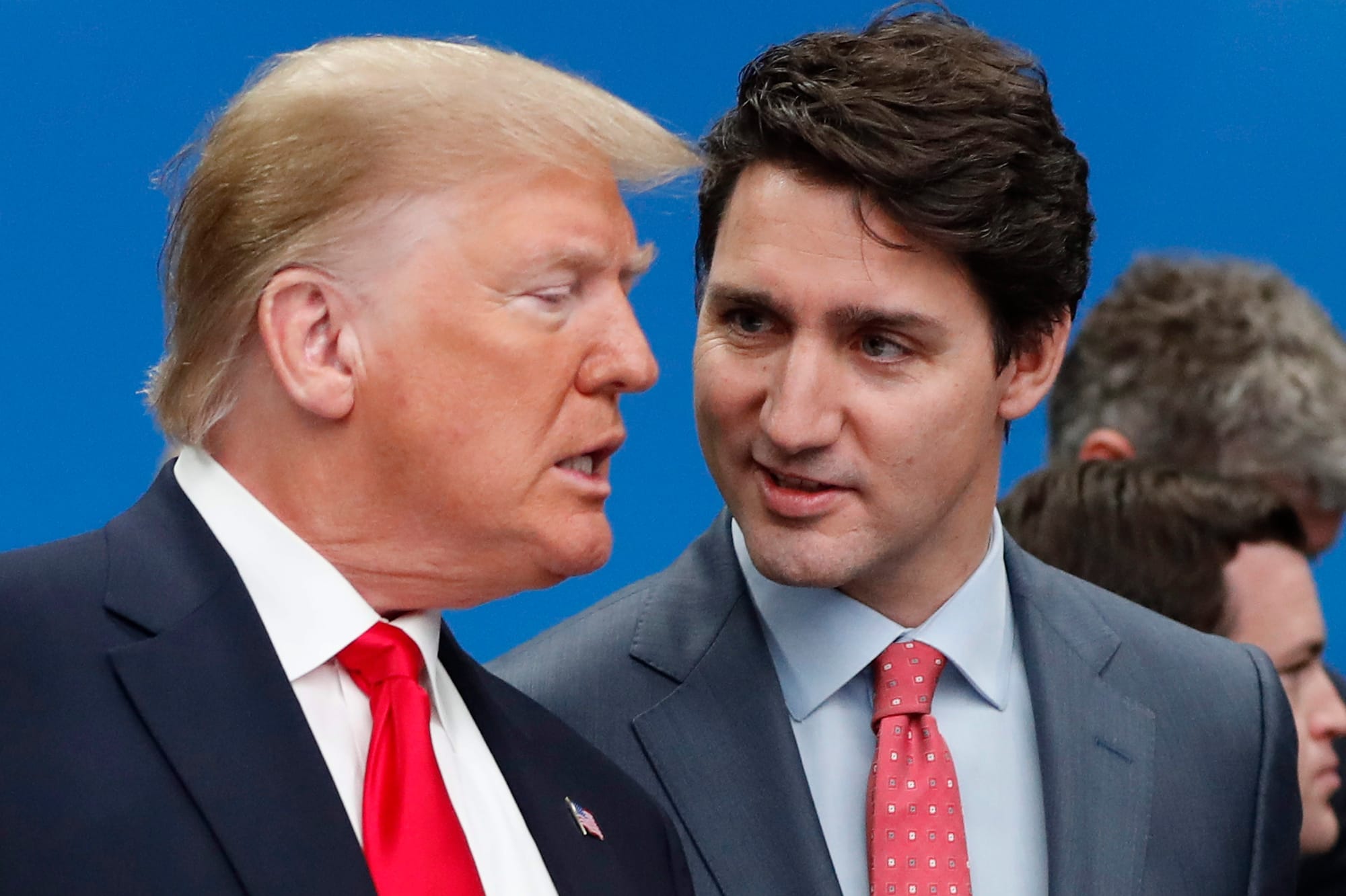 Side profile of Donald Trump leaning toward Justin Trudeau, the two stand against a blue background.