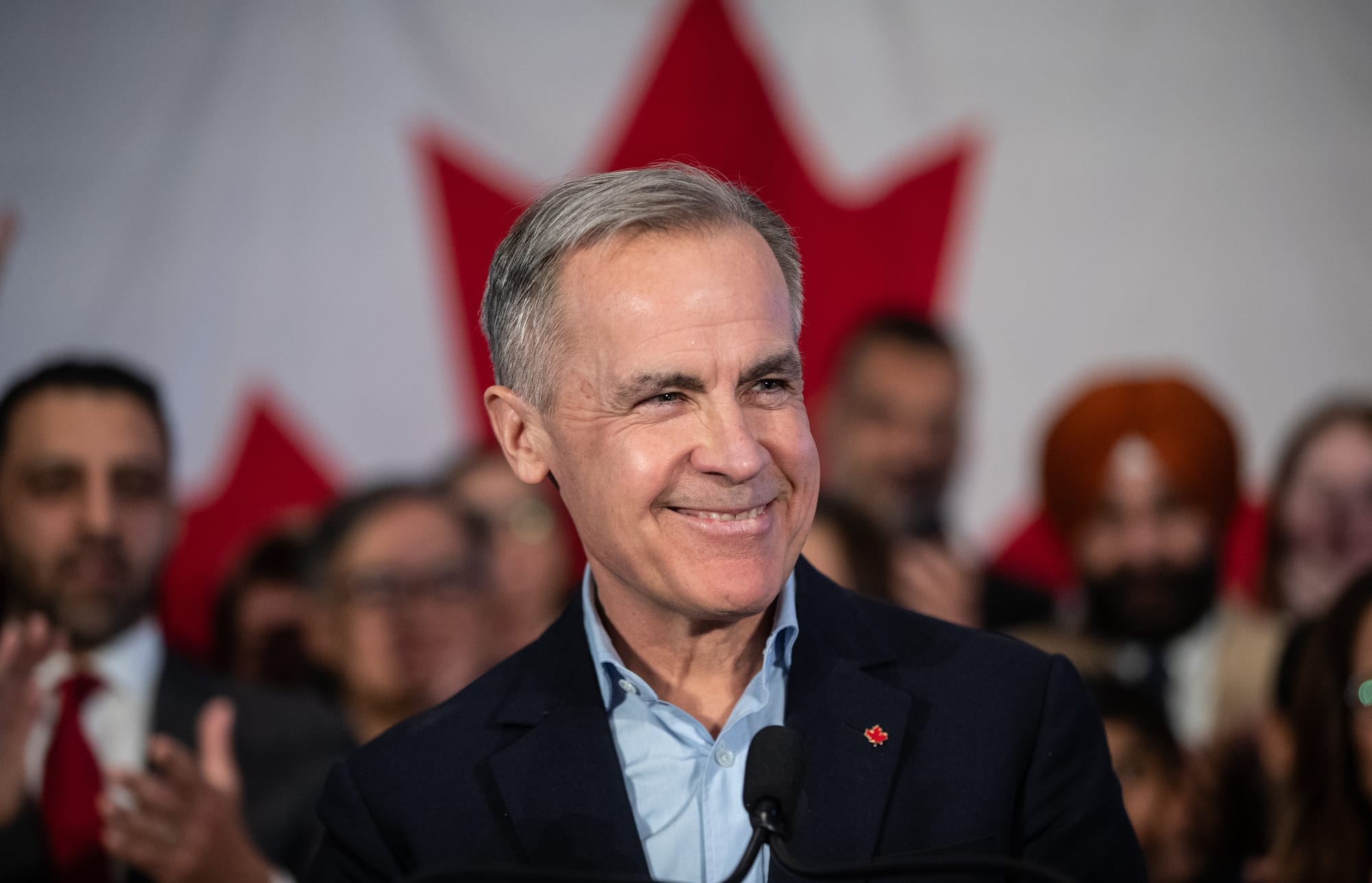 A man stands behind a mic speaking to a crowd. He wears a blue button up shirt under a black jacket with a maple leaf pin.