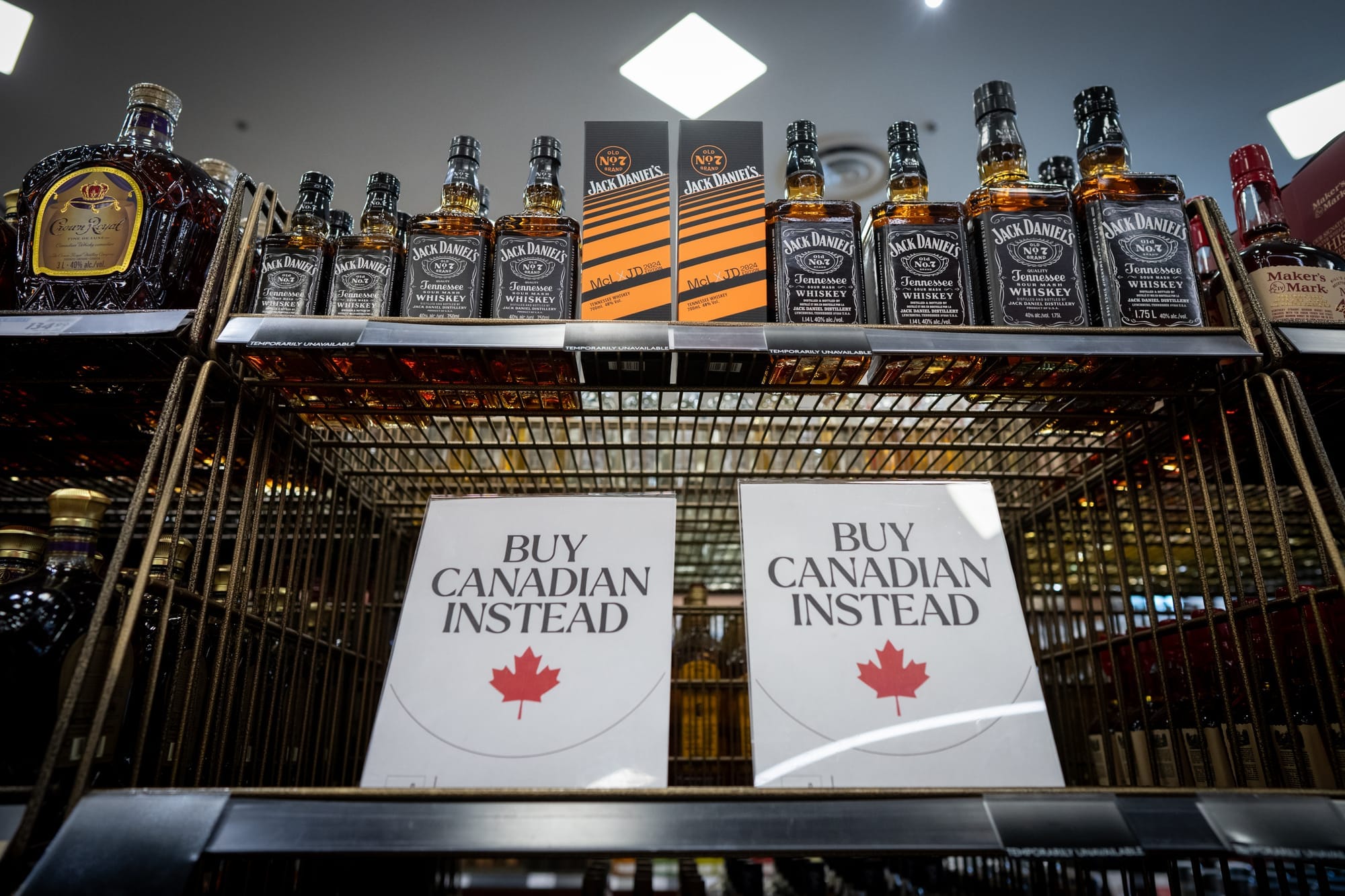 Two signs are placed on a liquor store shelf that say "BUY CANADIAN INSTEAD" with a maple leaf.