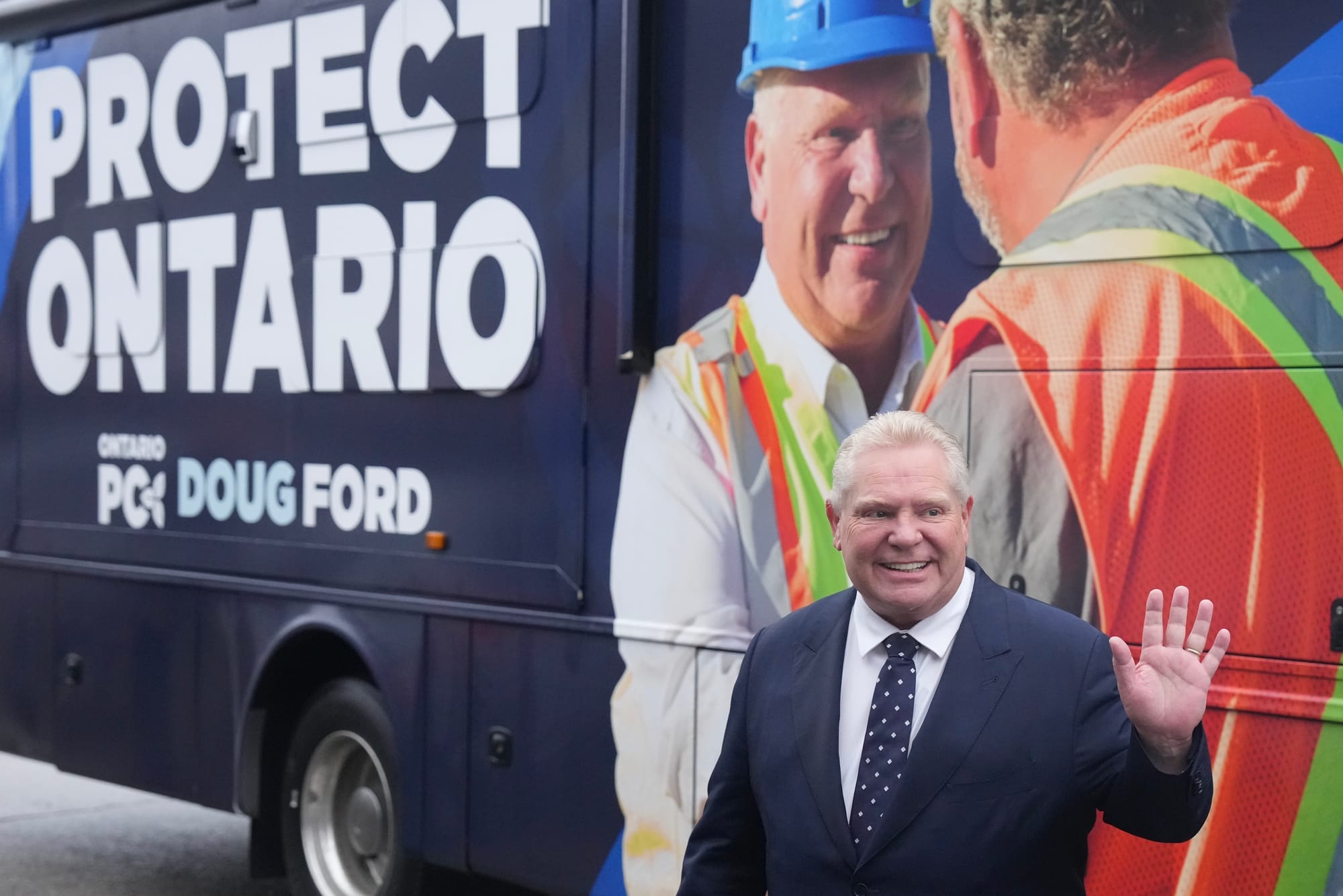 Premier Doug Ford smiles and waves into the distance as he stands in front of a campaign bus.