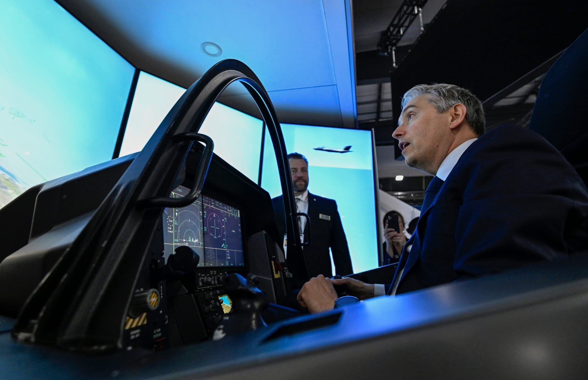 A man in a suit sits at a control panel for a fighter jet simulator