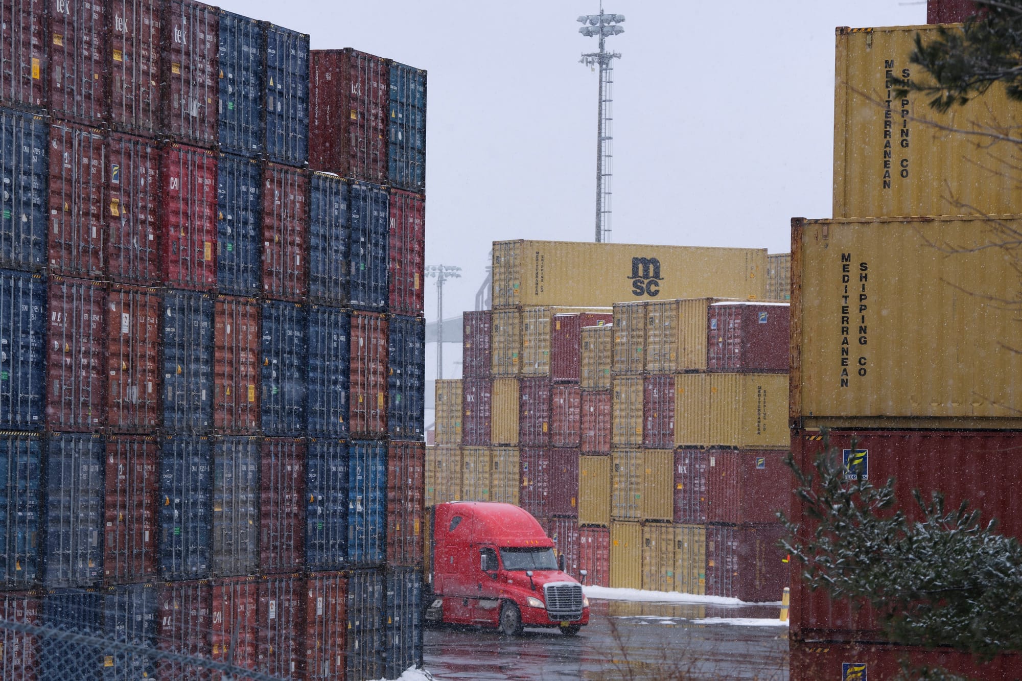 A red truck drives through stacked shipping containers at a port as it snows. 