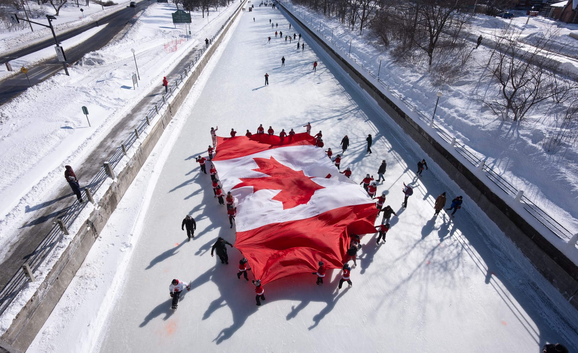 Overhead view of a group of young people holding a large Canadian flag as they skate on a frozen canal.