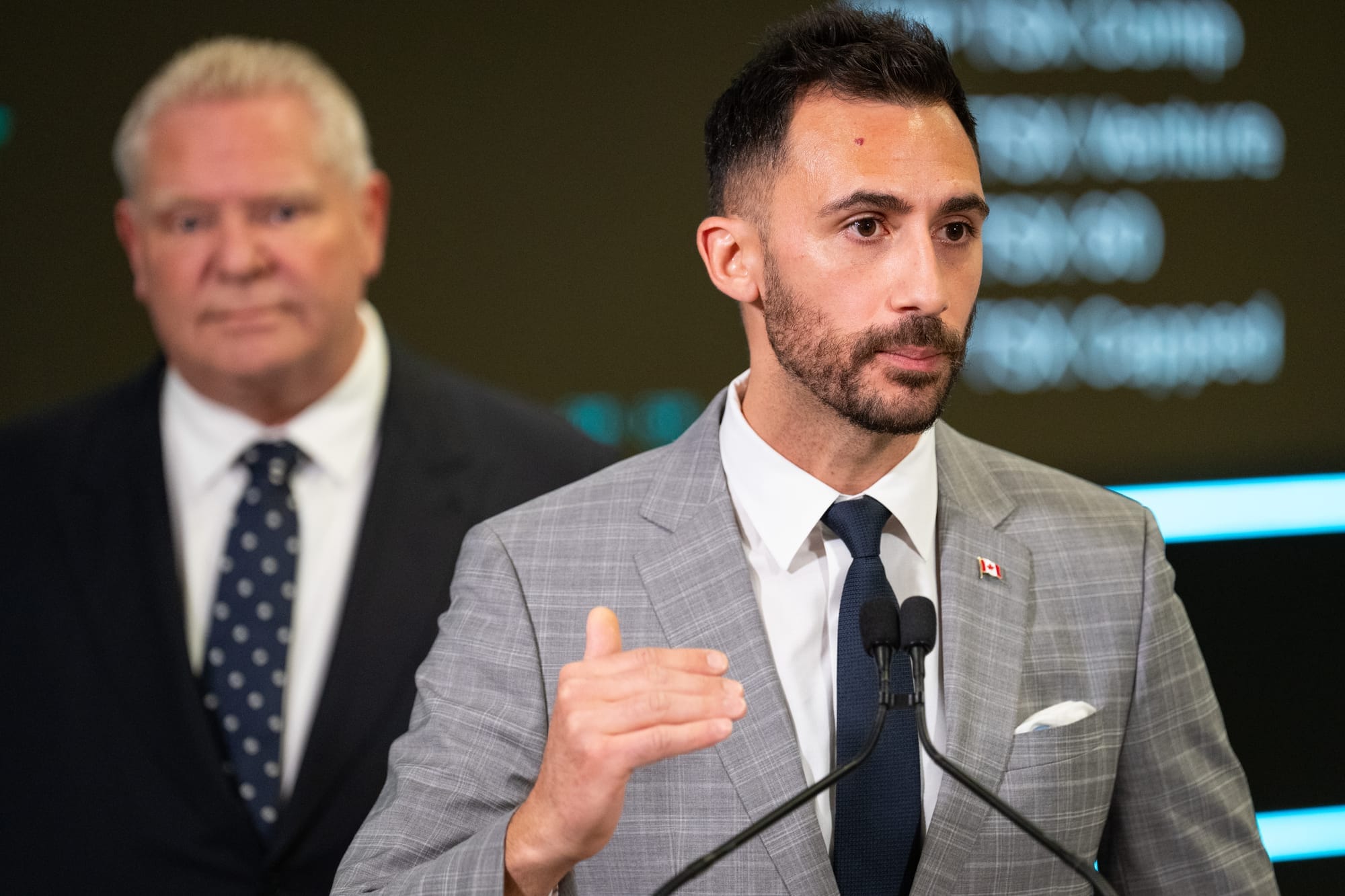 A man in a grey suit speaks at a press conference. Doug Ford is seen out of focus in the background 