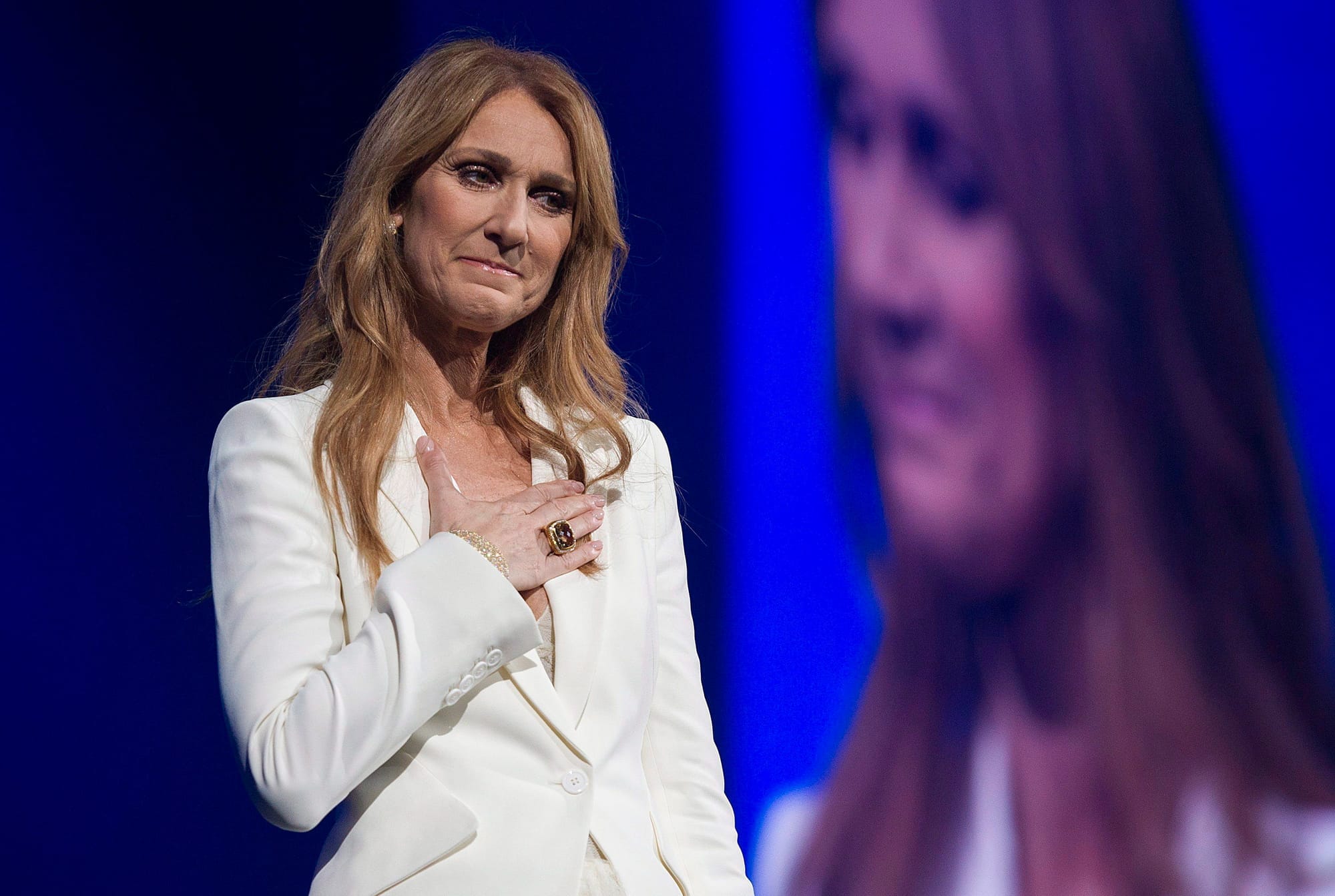 Celin Dion wears a white blazer and places her hand on her chest while performing in Montreal.