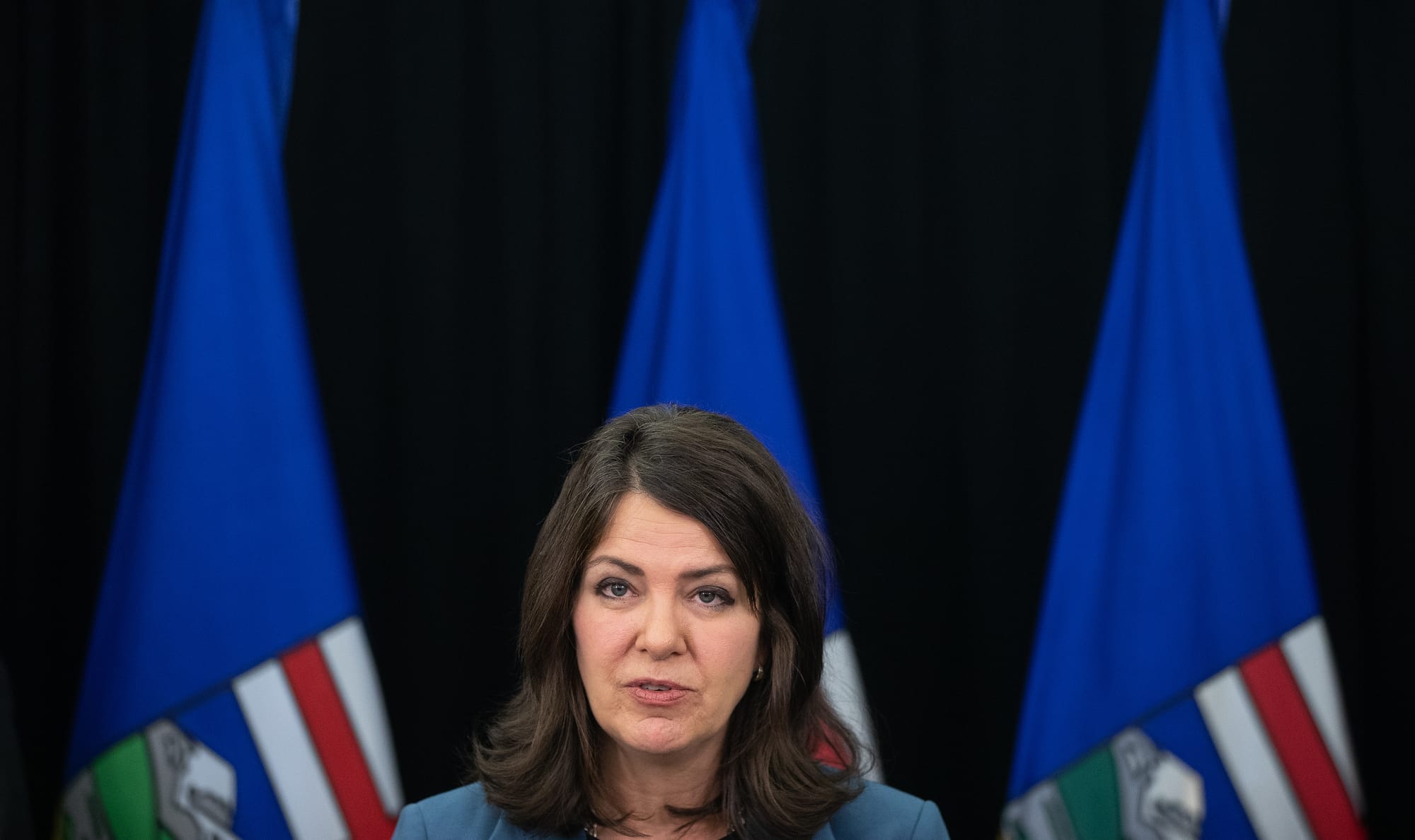Danielle Smith sits at a press conference in front of three Alberta flags