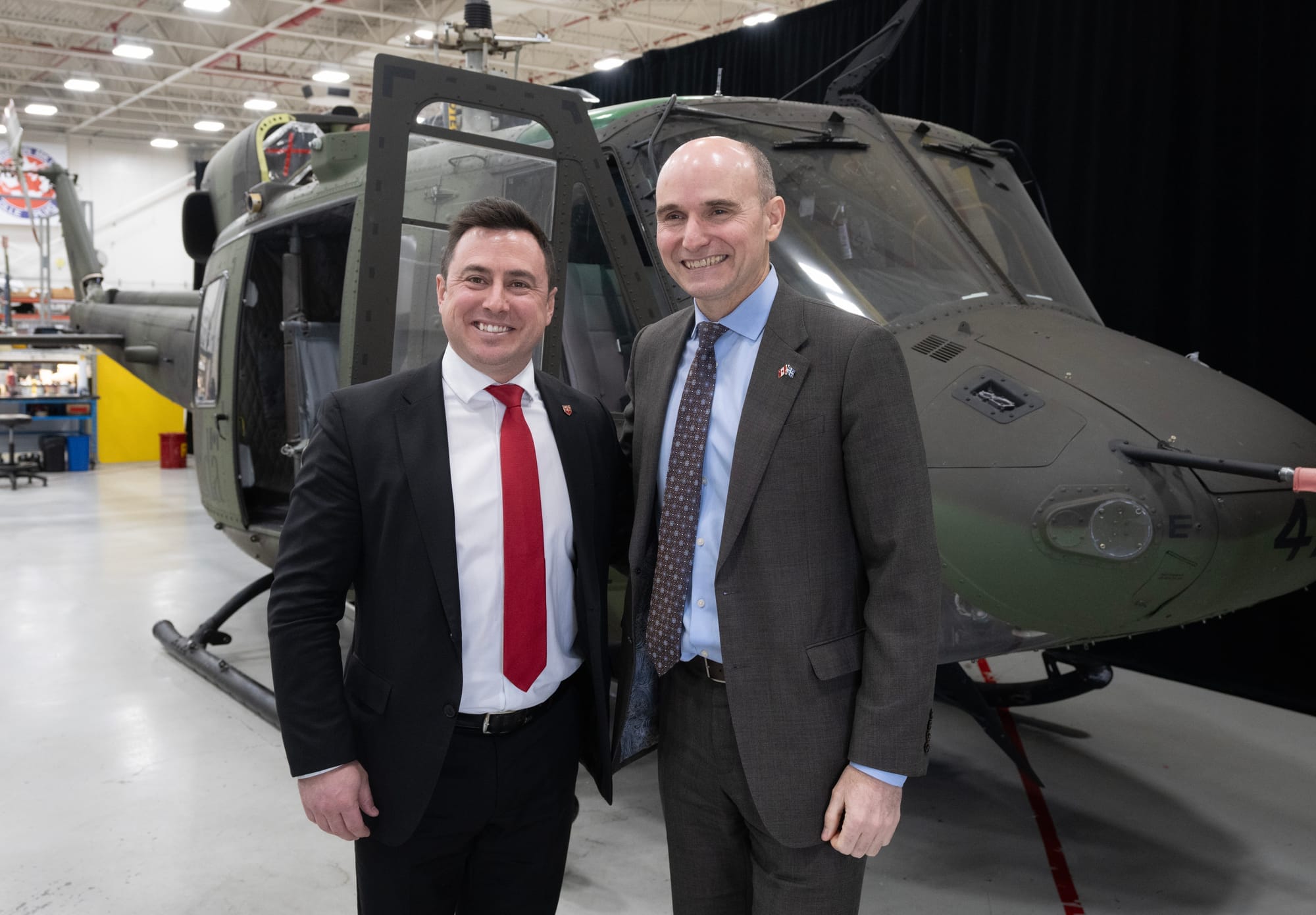 Two men in suits and ties stand in a warehouse in front of a helicopt