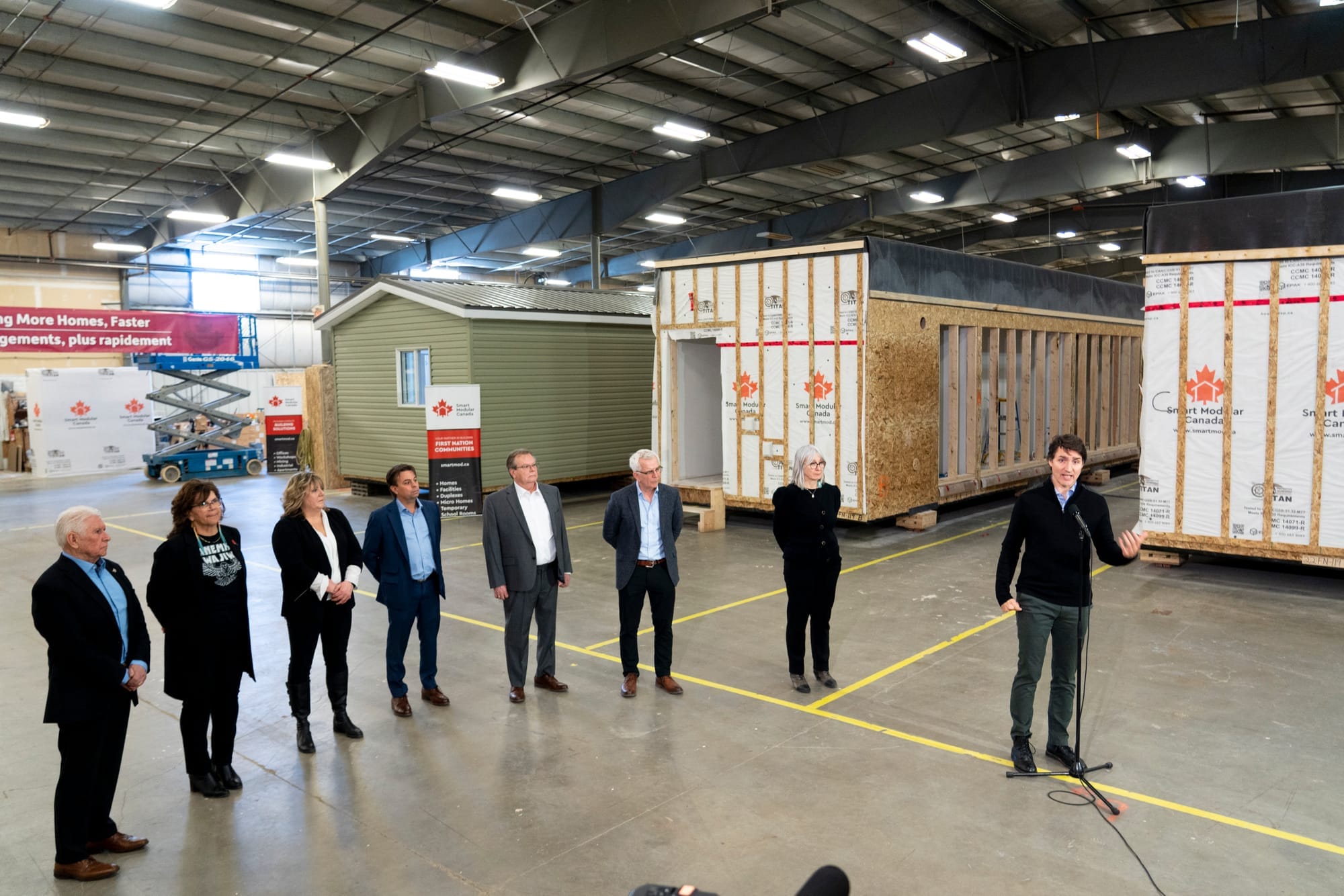 Justin Trudeau speaks in a warehouse surrounded by partially complete modular homes. Seven people look on.