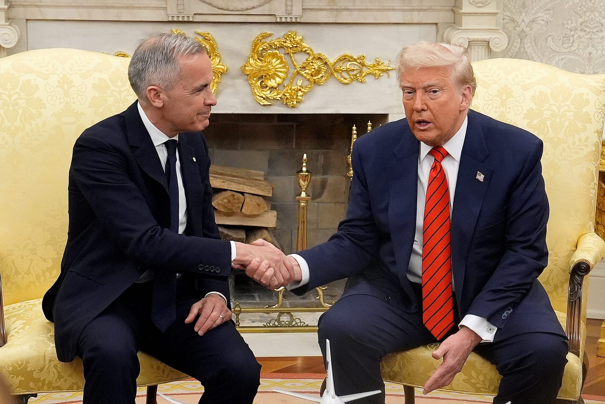 Mark Carney shakes Donald Trump's hand in two armchairs in front of an ornate fireplace. 