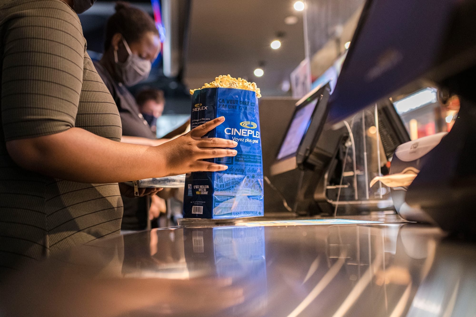 A person in a brown striped shirt holds a blue bag of popcorn with the Cineplex logo on it