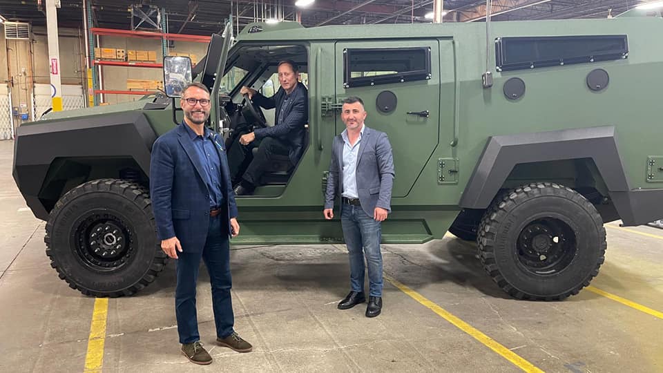 A man in a suit sits in a armoured truck with the driver door open, while two other suited men stand just outside of the vehicle