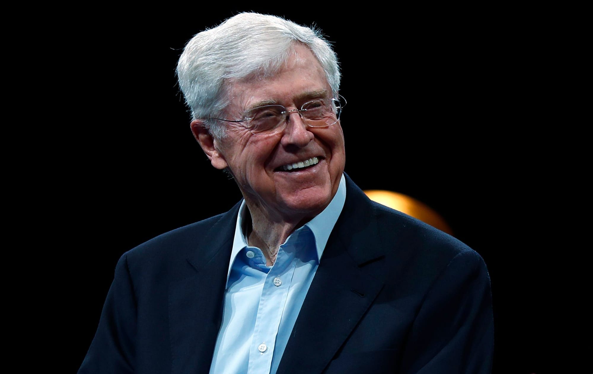A man with white hair and glasses smiles. He wears a button up shirt and suit jacket