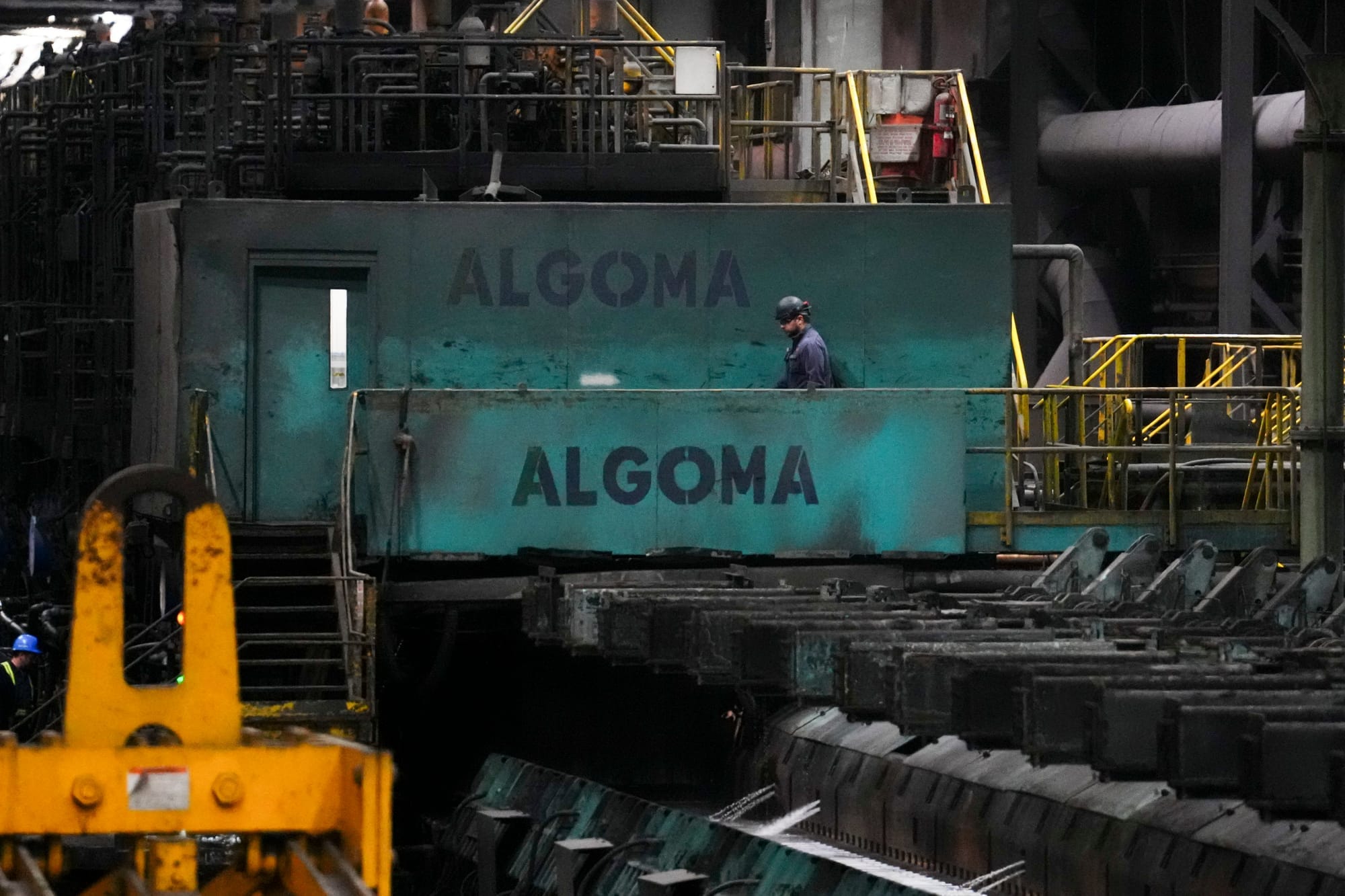 A worker in a hard hat walks past heavy machinery in a steel plant. The walls say ALGOMA.