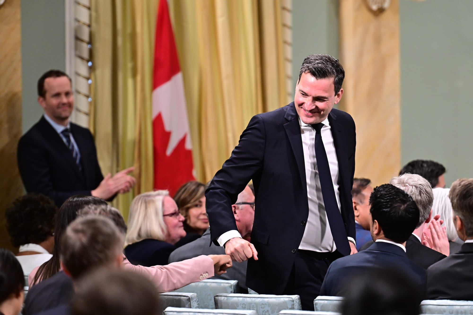 A man in a suit and black tie fist bumps a colleague as he stands and heads to the front of the room