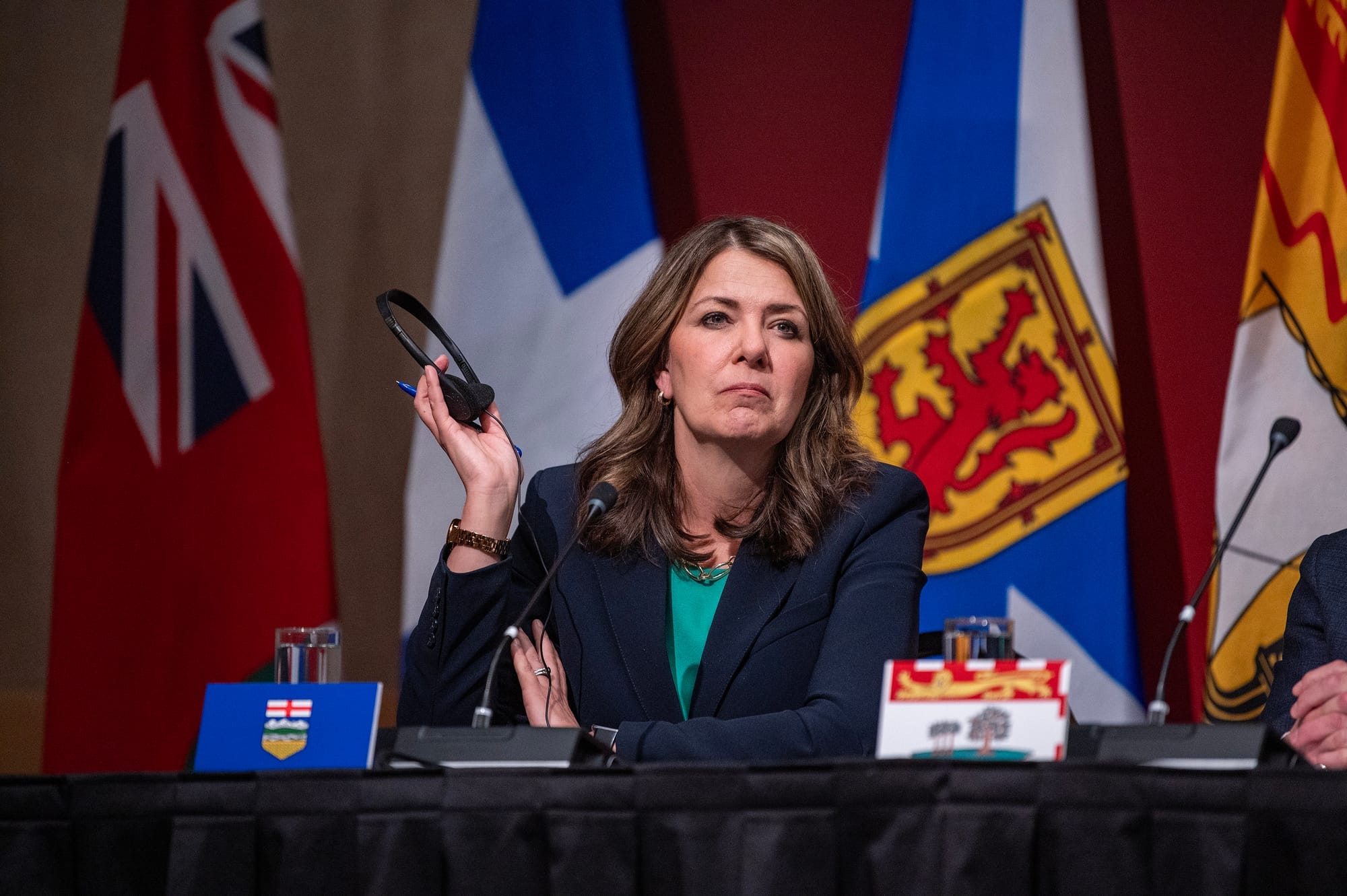 Premier Danielle Smith sits at a desk holding headphones, with flags in the background.