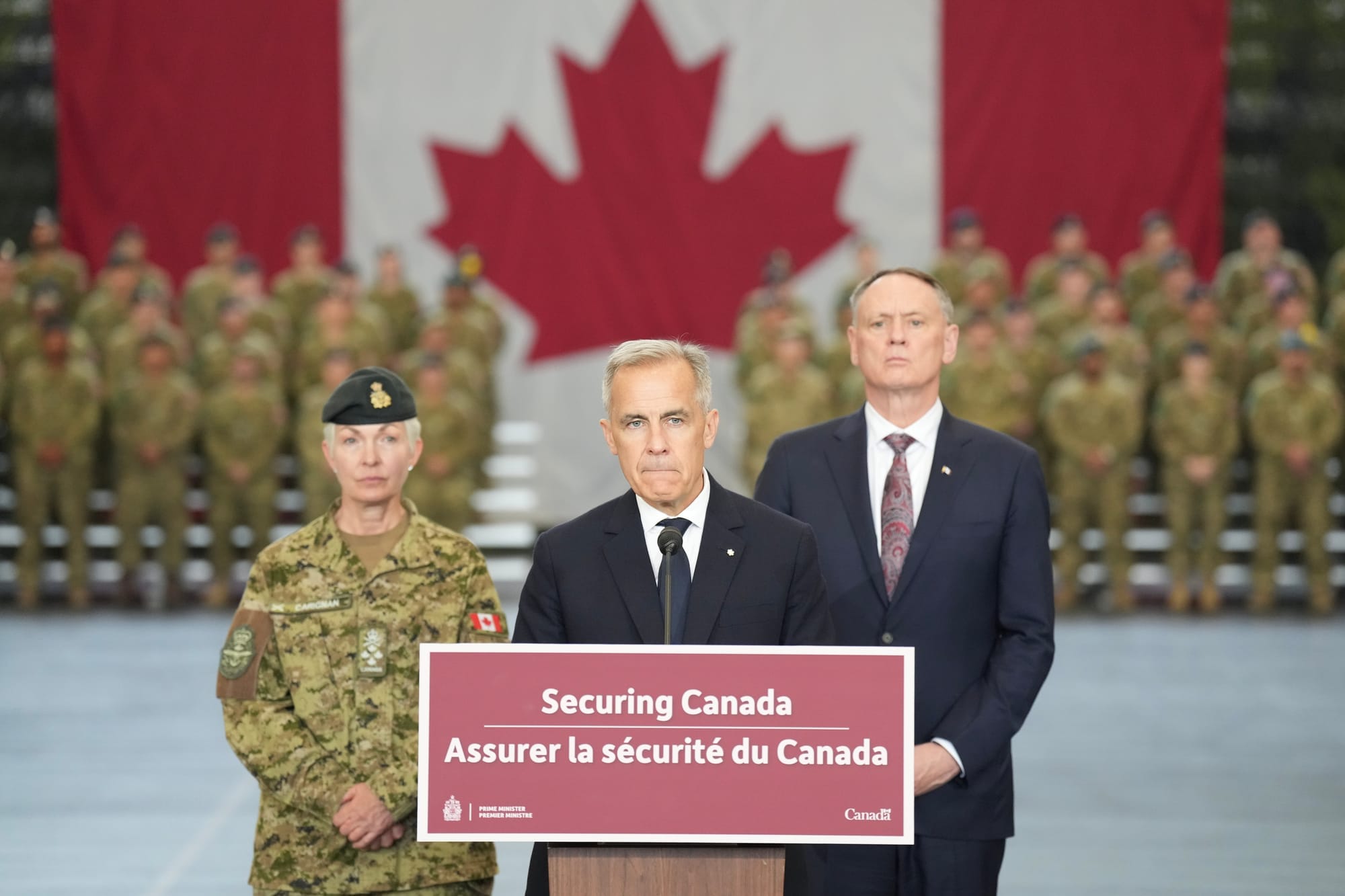 Two men in suits and a woman in military uniform stand behind a podium that says "securing Canada", in front of a Canadian flag and dozens of soldiers