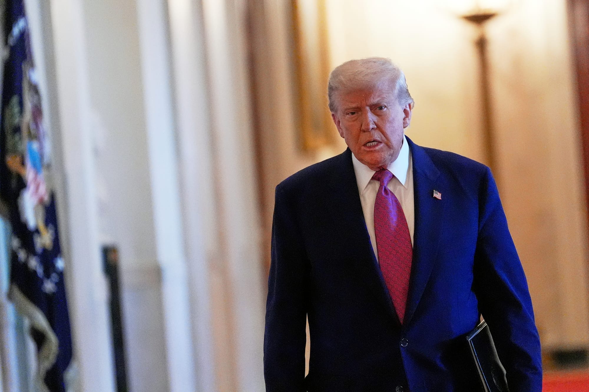 Donald Trump wears a blue suit and red tie and holds a folder of documents under his arm as he walks through the White Hou 