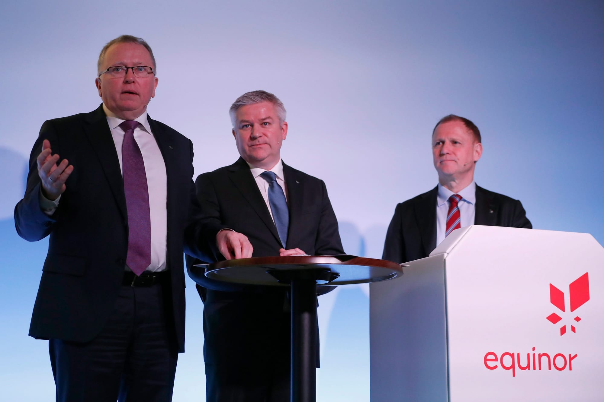 Three men in suits speak to a crowd. One stands behind a podium that says equin
