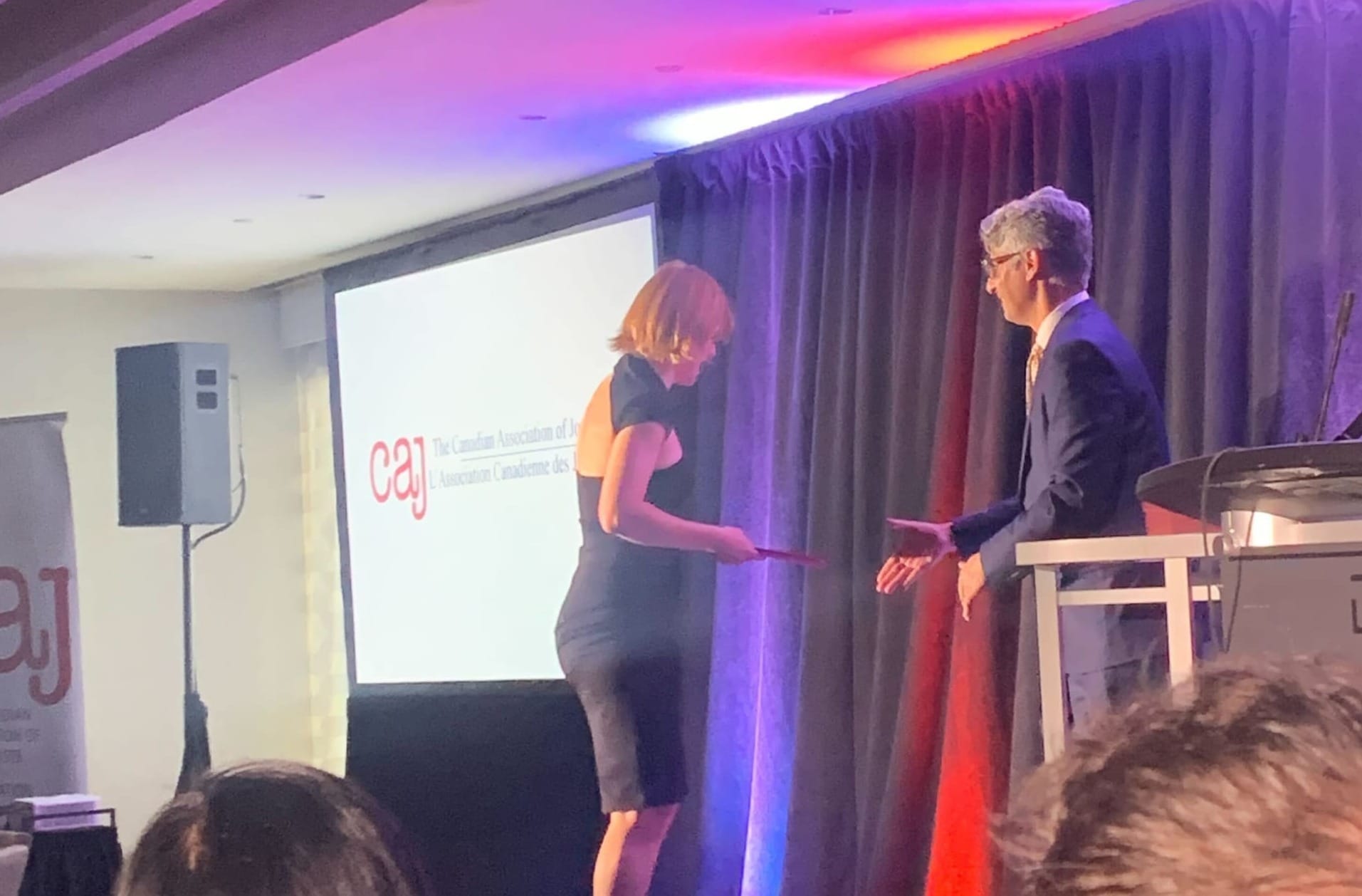 A woman in a black dress walks to the stage to accept an award from a man in a suit 