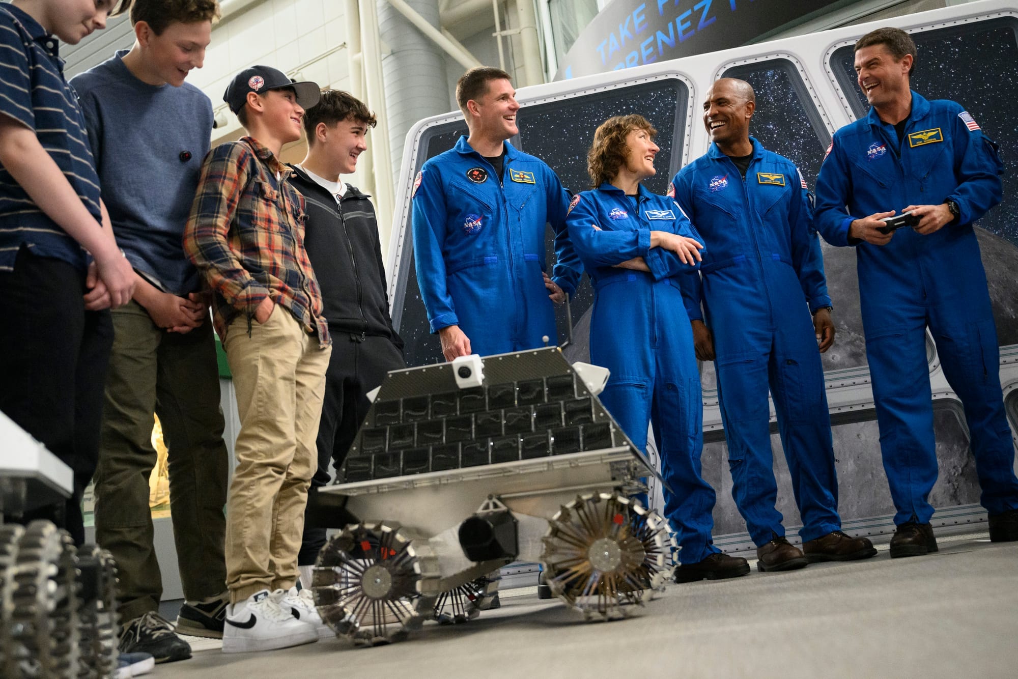 Astronauts in blue jumpsuits show a smal lunar rover to a group of students