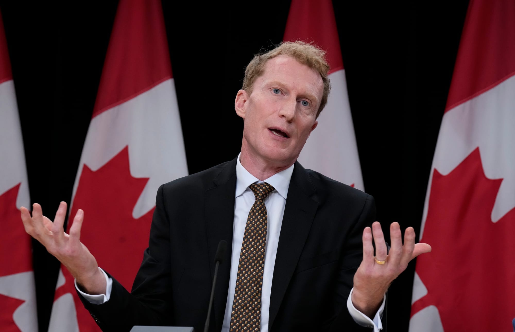 Marc Miller, seated in front of four Canadian flags, wears a suit and gestures as he speaks to reporters.