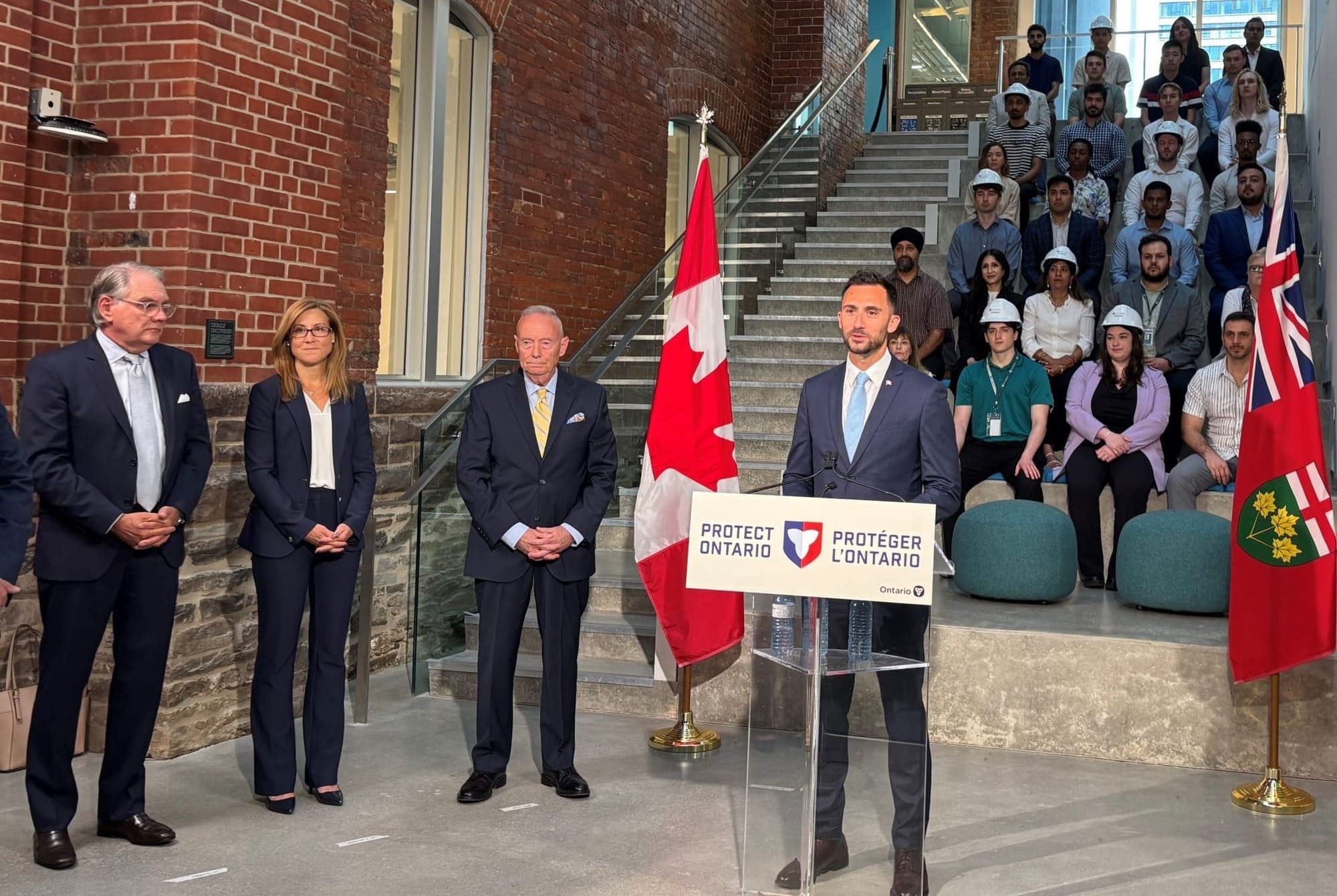 Energy and Mines Minister Stephen Lecce speaks to a group of GE Hitachi employees from a podium that reads "PROTECT ONTARIO" 