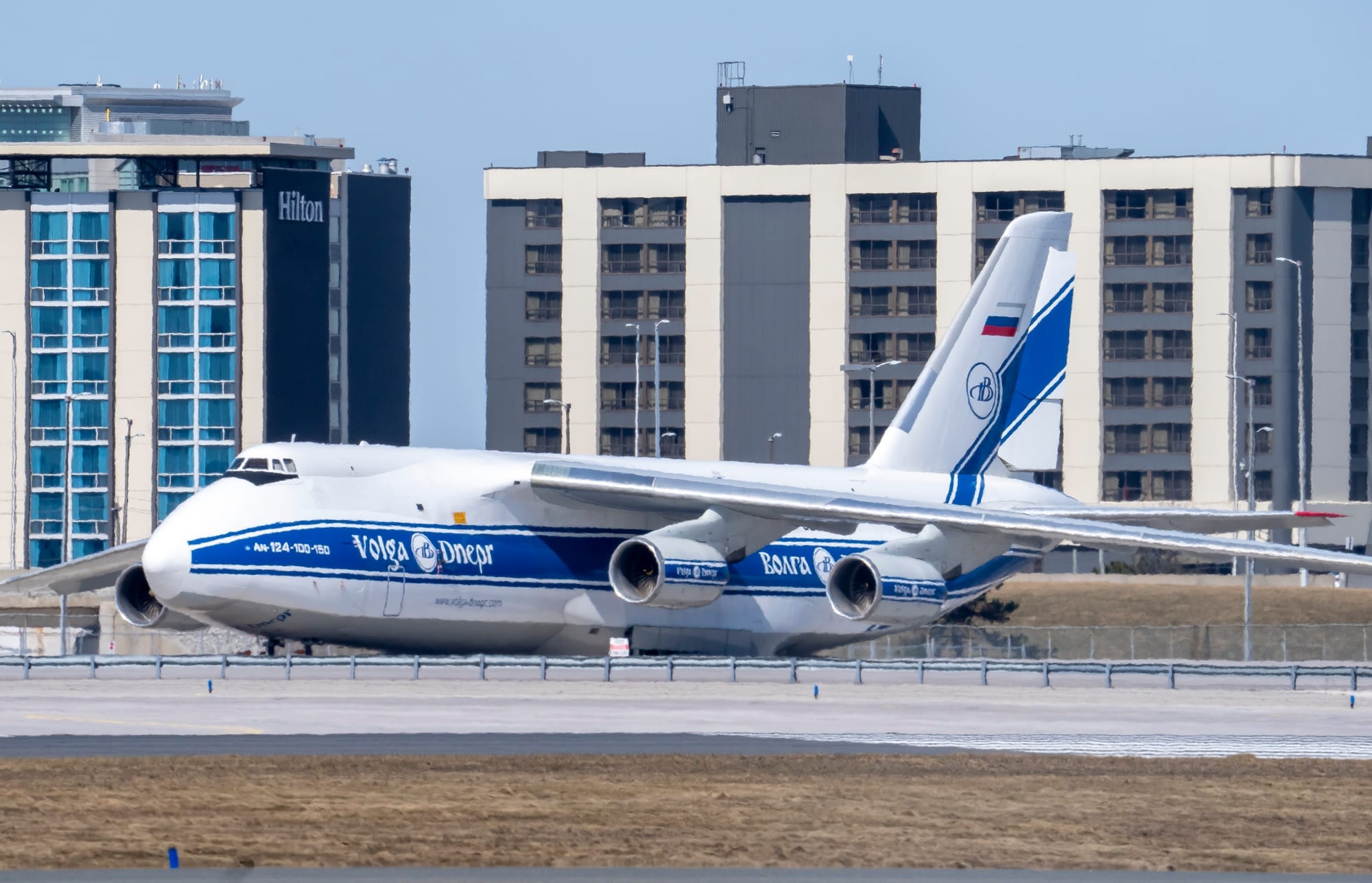 A white and blue cargo plane sits on airport tarmac. A white logo on the side reads "Volga Dnepr."