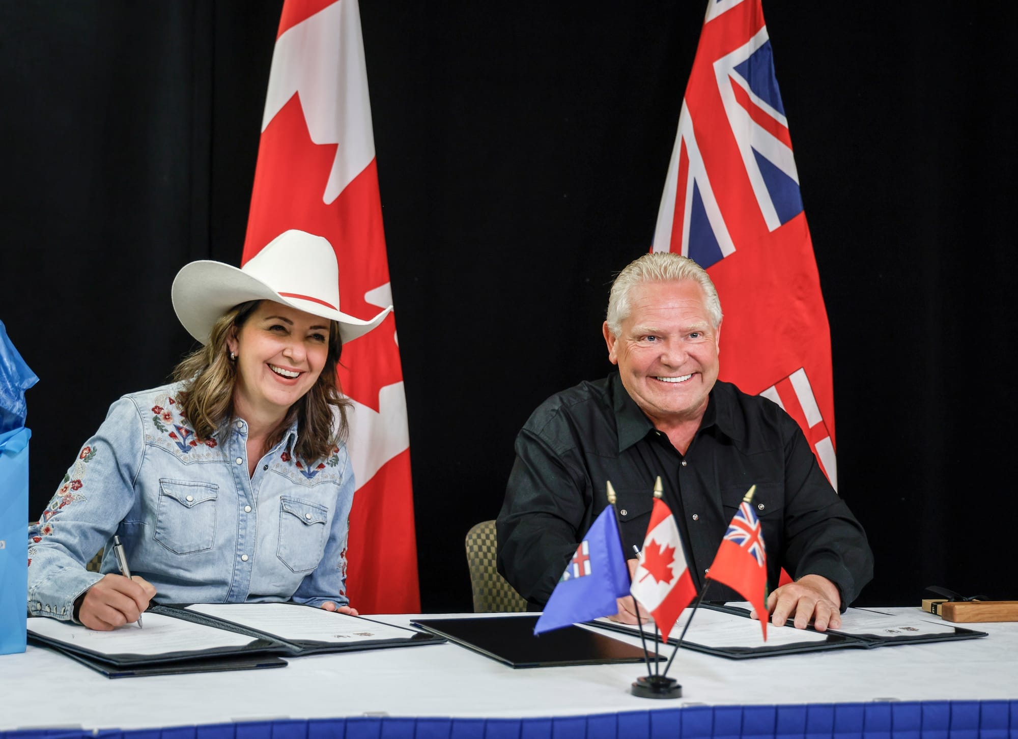 Doug Ford and Danielle Smith sign agreements sitting at a table with the Ontario, Alberta and Canadian flags.