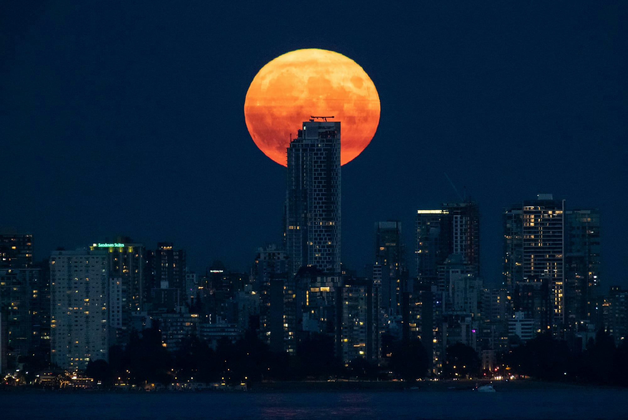 A bright orange full moon rises behind a condo tower in a nighttime photo of the downtown Vancouver skyline.