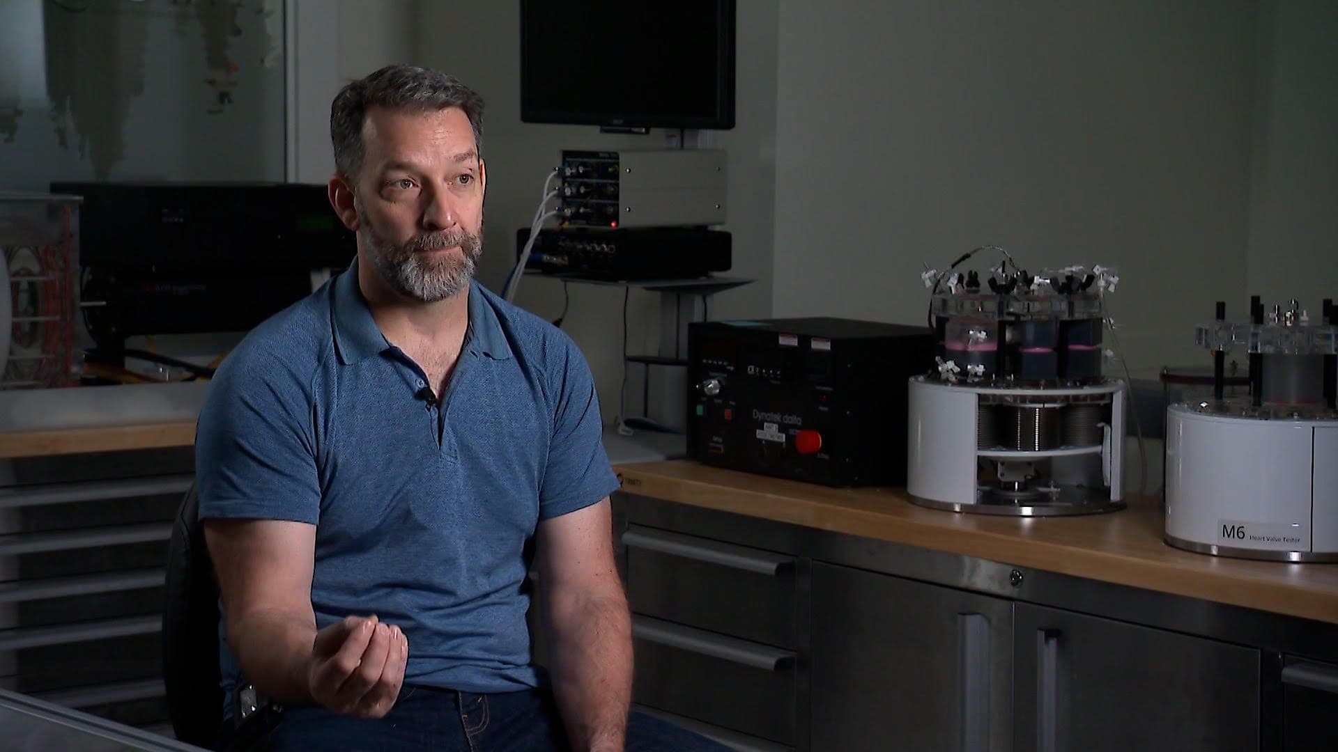 A bearded man wearing a blue shirt sits in the corner of a laboratory.